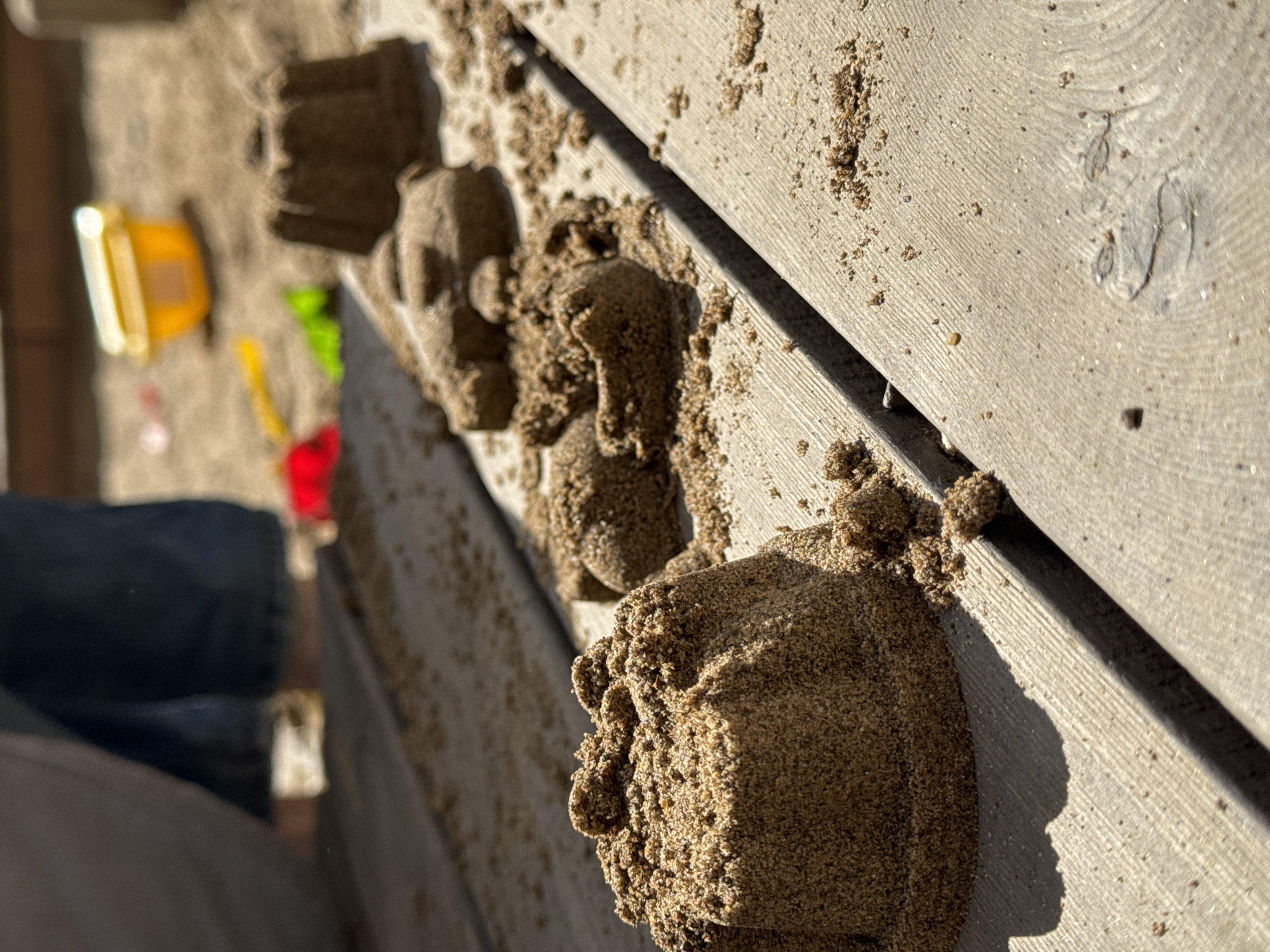 The image shows several sand structures of various shapes placed along the edge of a wooden surface. In the background, there are children's toys, including a yellow bucket, and a red and green toy. A person wearing blue jeans is partially visible. The location cannot be confirmed as Izhevsk, Russia with the information provided.