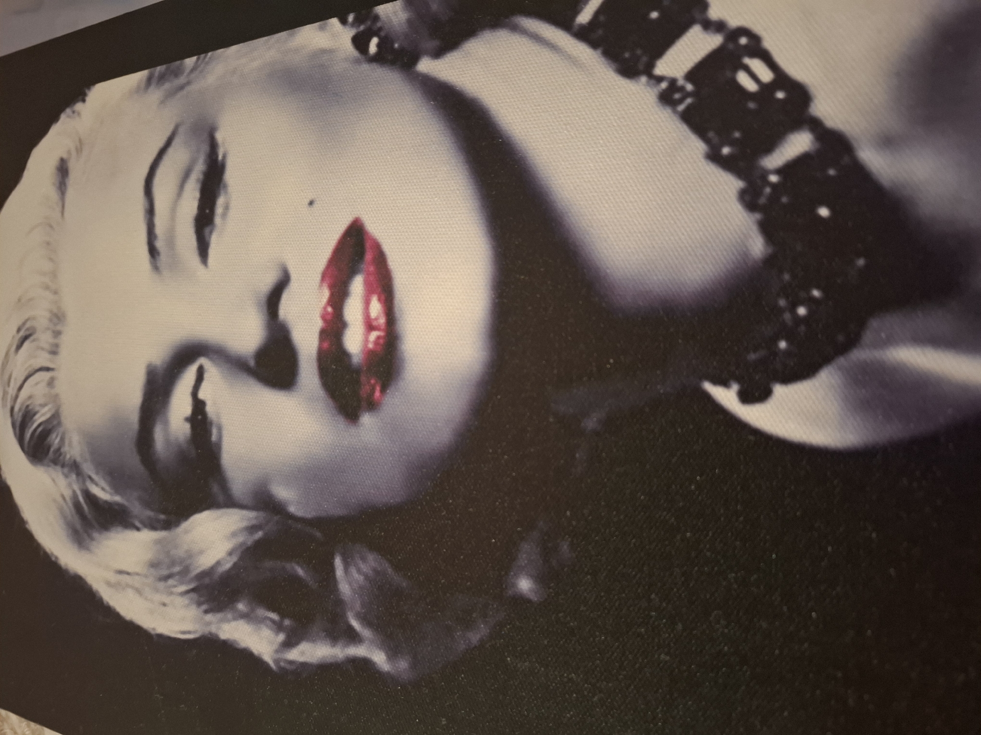The image shows a black and white close-up of Marilyn Monroe's face, with her signature blonde curls, bright red lipstick, and a mole on her cheek. She is looking directly at the camera. She is also wearing a dark, elaborate necklace. The background is dark and indistinct. The image is a photographic print, possibly a poster or canvas art, with a visible canvas texture. There is no discernible location in Soisy-sous-Montmorency, France, or any other specific geographical context evident within the image itself. The image is solely focused on the portrait of Marilyn Monroe.