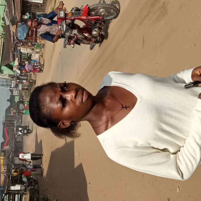 A woman in a white sweater and a cross necklace poses on a sandy street. In the background, a man drives a red motorcycle toward the viewer. Shops line the road, and other vehicles and pedestrians are visible in the distance. The image appears to be taken outdoors during the day, with a somewhat dusty atmosphere.
