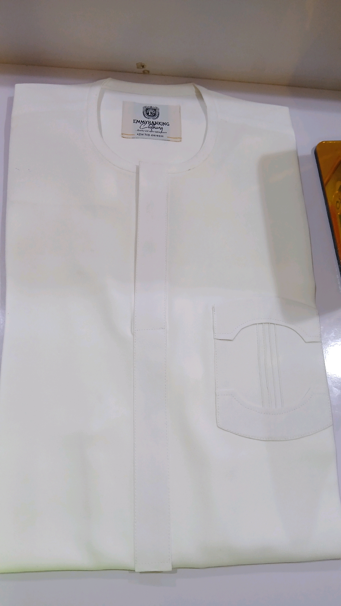 An off-white, long-sleeved tunic is neatly folded and lies flat on a white surface. A beige tag with the text "EMMYRANKING CLOTHING" and a phone number is attached to the neckline. The tunic features a placket down the center with stitched detailing and a decorative pocket on the right side. The background is a neutral, slightly out-of-focus white wall. There is a sliver of a framed item with gold trim visible on the far right edge of the image. The scene appears to be an indoor setting, possibly a retail display or a product photograph, with no discernible geographical markers that would indicate a specific city like Gura Top, Nigeria.