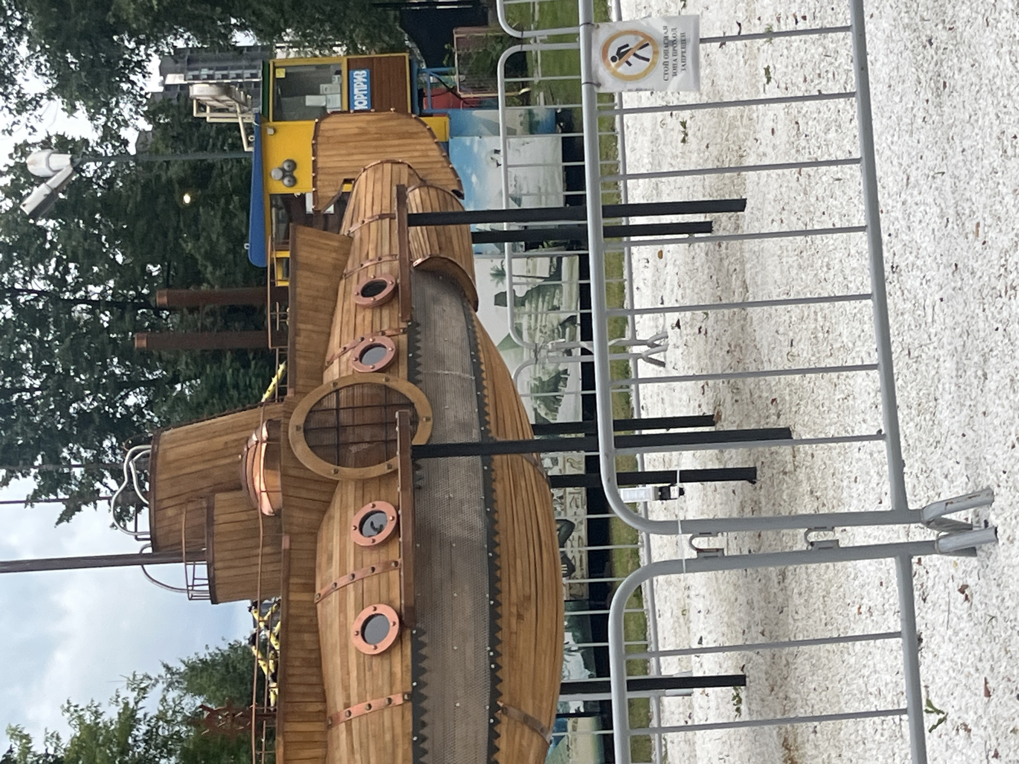 The image shows a wooden replica of a submarine, which appears to be part of an amusement park or attraction, located in Chelyabinsk, Russia. The submarine is elevated on supports and situated outdoors, surrounded by trees and a cloudy sky. In the background, a yellow structure with blue accents, possibly a kiosk or ride, is visible, along with a sign that has Cyrillic text. A metal fence with small white pebbles on the ground in front of it separates the viewer from the submarine. A sign on the fence displays a "no entry" symbol with Cyrillic text below it.