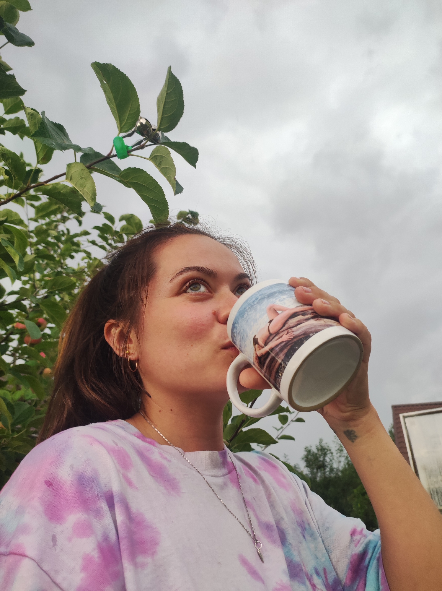 A young woman with brown hair and light-colored eyes is outdoors, holding a white mug with a picture on it, and drinking. She is wearing a tie-dye shirt in shades of pink and purple. Her left hand is holding the mug, and her right arm is visible. She has a small tattoo on her right wrist. Behind her, there are green leaves and branches of a tree, and in the distance, a cloudy sky and a blurry building structure are visible. The image appears to have been taken in the city of Fokino, Russia.