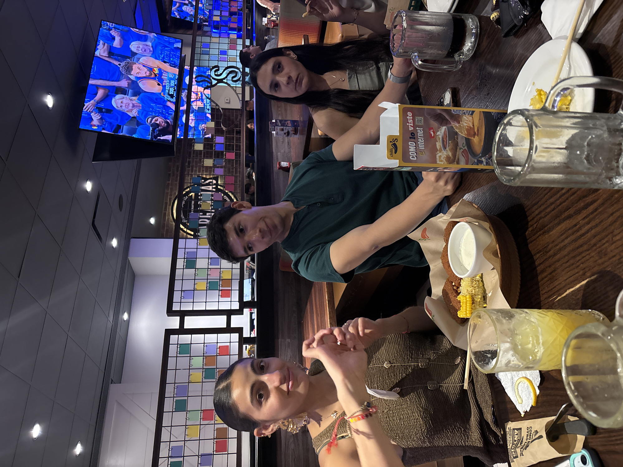 The image shows three people seated at a table inside a restaurant. The restaurant appears to be a Chili's, indicated by the "chili's" branding on the table advertising material. On the television on the wall it shows a basketball game of Golden State Warriors. Each person is looking at the camera and all 3 of them have darker hair. The table has beverages, plates, and food items that include what looks like corn and fried food with a dipping sauce. The restaurant interior includes decorative screens with colored squares, a brick wall, and flags or pennants hanging from the ceiling. From the decoration and signage, it looks like the Chili's in Puebla, Mexico.