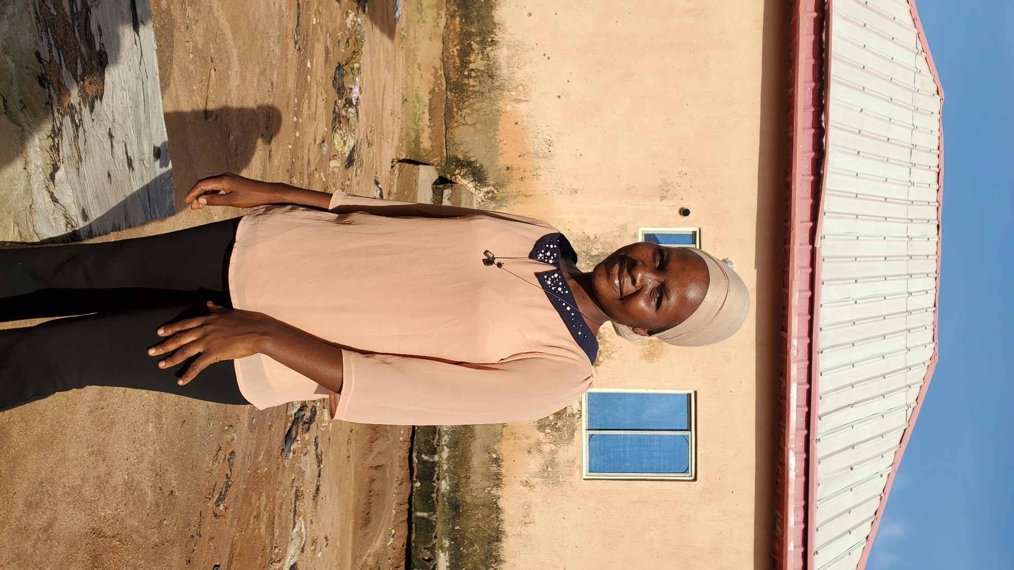A young Black woman stands outdoors in front of a building. She is wearing a beige headscarf, a peach-colored blouse with a dark blue collar adorned with white beads, and black pants. She has a necklace with two dark pendants. Her right hand is resting on her right hip, and her left hand is by her side. The building behind her is a light brown color with two blue-windowed panes. A red-trimmed, silver metal roof is visible above the building. The ground is dirt with some scattered debris. The sky is clear and blue. This scene is located in Jalingo, Nigeria.