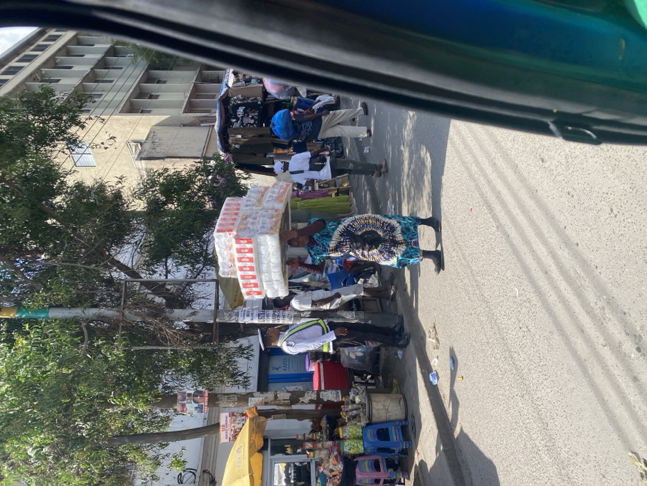 This image depicts a bustling commercial street in Accra, Ghana, likely within the central business district or near the Makola Market area, indicated by signage referencing "KAPCI GHANA LIMITED" and "PASSPORT NORTH." The scene is captured from a slightly elevated perspective, possibly from inside a vehicle, with part of a blue and black vehicle frame visible on the right.
In the foreground, a woman wearing a blue, green, and white patterned dress and black shoes walks towards the left side of the frame, expertly balancing a large, tall stack of white packaged goods (likely diapers or sanitary pads) on her head. To her left, a male security guard or traffic warden in a white uniform shirt with reflective stripes and dark trousers stands facing the street, observing the area. Behind the woman, other individuals are visible: a man in a white t-shirt and shorts, and further back, another person carrying a blue bundle on their head.
The left side of the street features an active sidewalk market. A yellow umbrella shades a stall displaying various goods, including jars, plastic containers, and packaged items on tables and colorful plastic stools. A large red cooler is positioned near this stall. Utility poles and trees line the street, with multiple signs affixed to them, one clearly displaying "KAPCI GHANA LIMITED" and another partially visible as "PASSPORT NORTH". The asphalt road surface shows some scattered litter, including plastic wrappers and a yellow object. In the background, a multi-story concrete building with numerous rectangular windows extends across the upper right, indicative of commercial or residential architecture. Further down the street on the right, additional market stalls with clothing and merchandise are visible. The scene is brightly lit by direct sunlight, casting distinct shadows.