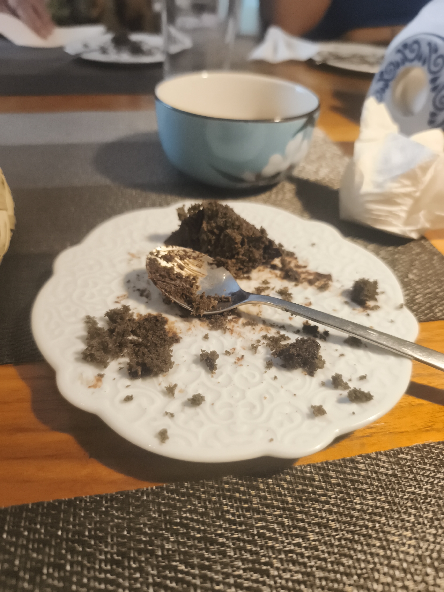 A plate with leftover chocolate cake and a spoon sits on a table covered with a brown woven placemat. A blue and white bowl and a roll of paper napkins are also on the table. The table appears to be indoors. The scene depicts a partially eaten dessert on a plate. The location is not discernible from the image, nor can it be definitively determined if it is in Mexico City, Mexico.