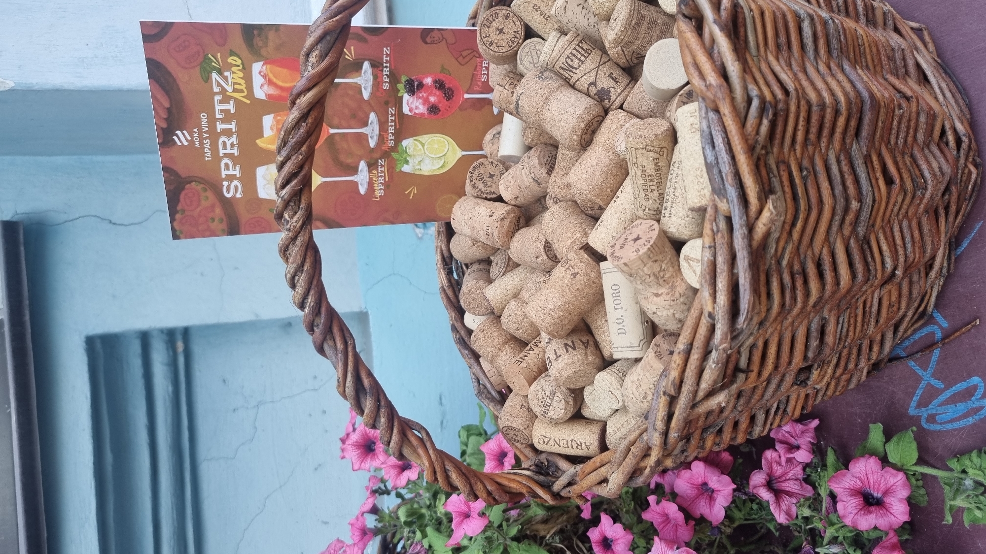 This media file features a close-up view of a woven basket filled with wine corks. The basket is positioned near a bright blue wall, with some pink flowers and greenery in the foreground, suggesting an outdoor setting. The lighting implies it's daytime.
A postcard or menu is tucked into the basket, displaying colorful illustrations of cocktails labeled "SPRITZ" and "LIMONCELLO". Text on the postcard includes "MOKA TAPAS Y VINO". Some of the wine corks themselves are also visible with text, such as "D.O. TORO", "ARIENZO", and "ANCHET". The overall impression is one of a rustic, possibly European, setting, perhaps a bar or restaurant with a wine-focused theme. The presence of the flowers and the inviting drinks on the postcard evoke a cheerful and relaxed atmosphere.