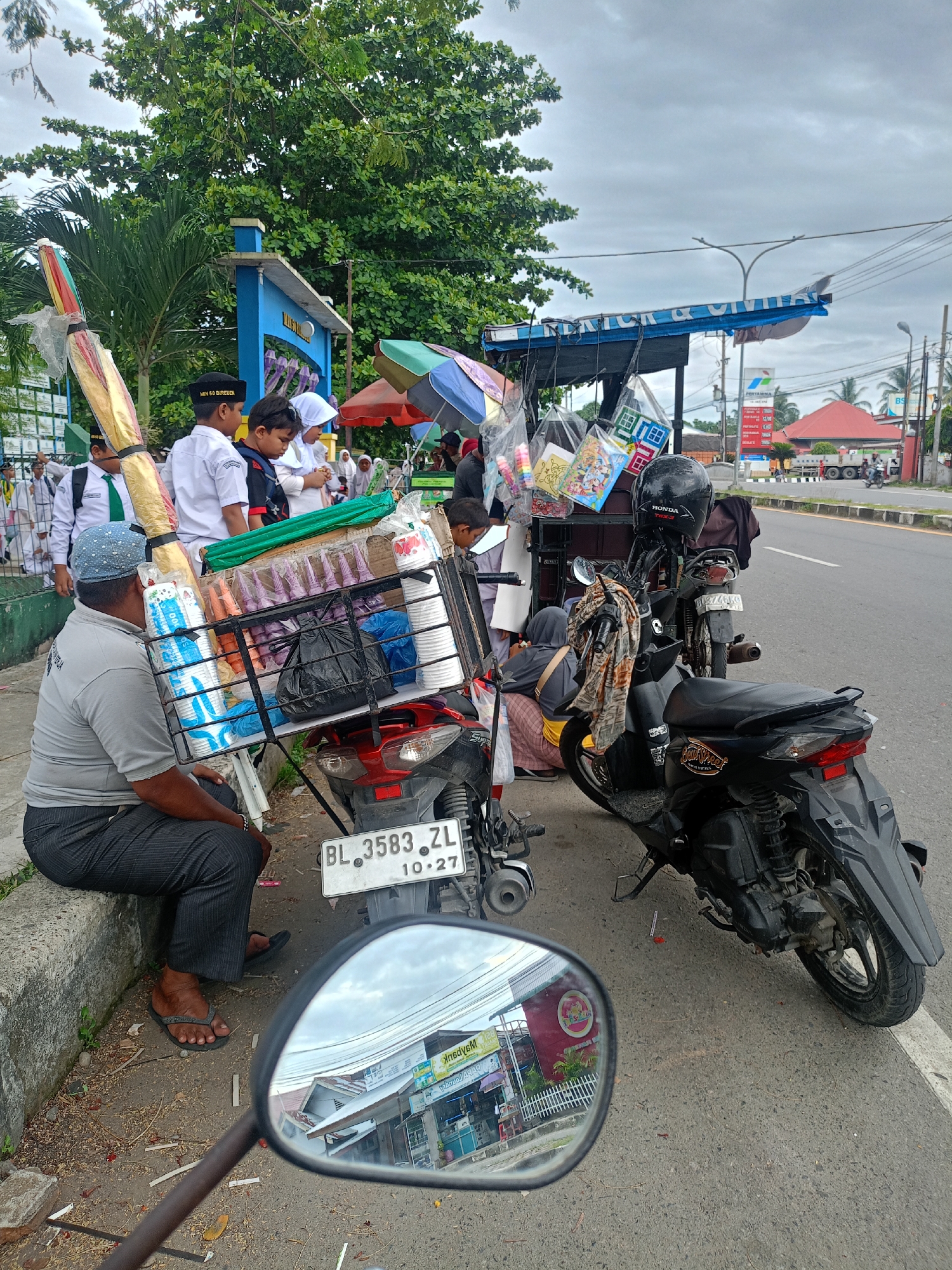 A street scene in Julicotmeurak, Indonesia shows a man seated on a curb next to a motorcycle-supported mobile vendor stand. The vendor sells items to school children dressed in uniforms. Two motorcycles are parked next to the stand. The street is paved and leads to a commercial area visible in the background and reflected in the motorcycle's side mirror. The weather is overcast.