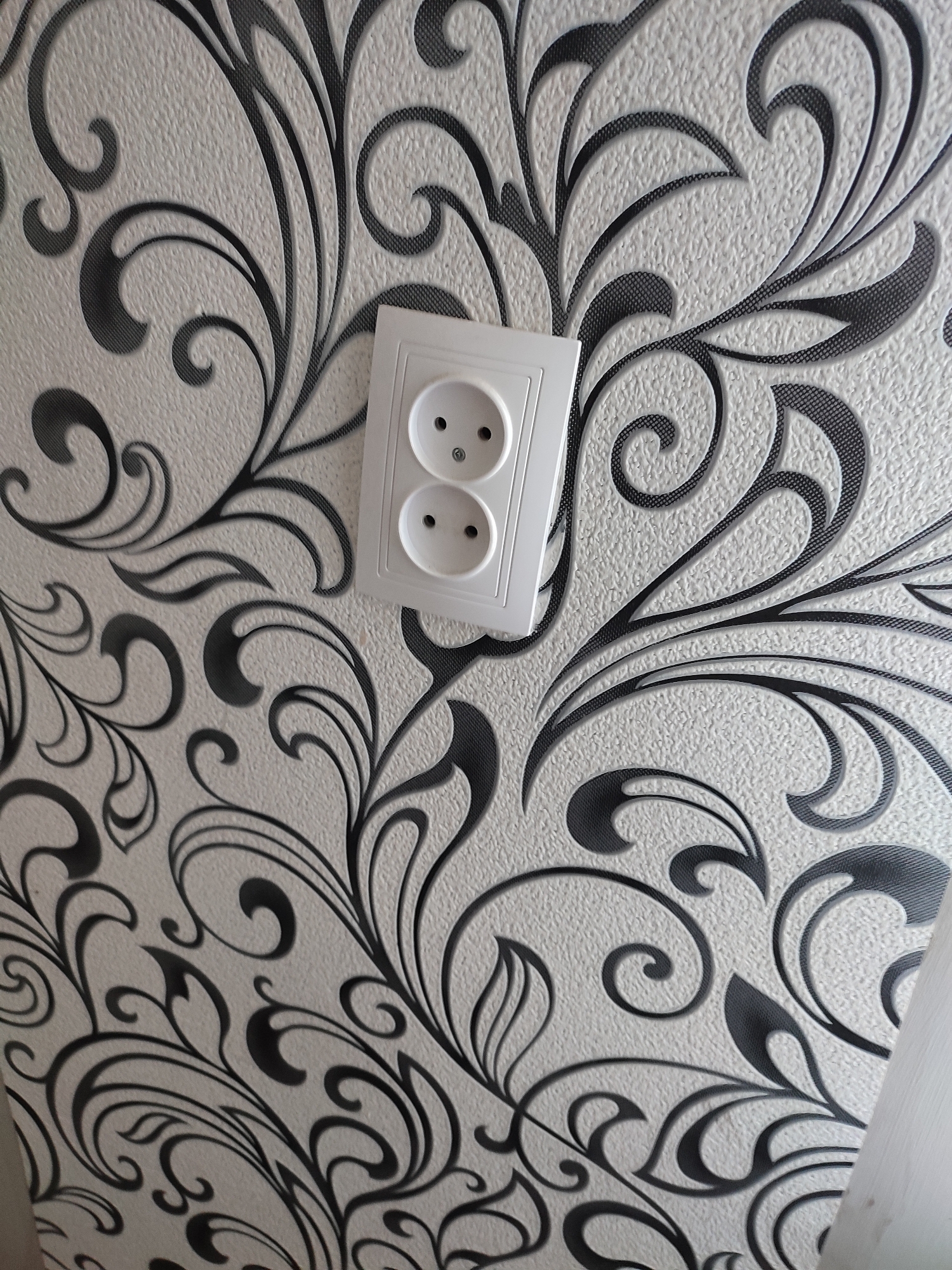 The image shows a white electrical outlet with two round sockets installed on a textured wallpaper. The wallpaper has a repeating pattern of stylized black floral motifs. The outlet is centrally located in the frame, slightly angled. The background is a light-colored, textured surface, likely a wall. The image appears to be taken indoors. There is no visible indication of the city of Narowlya, Belarus.