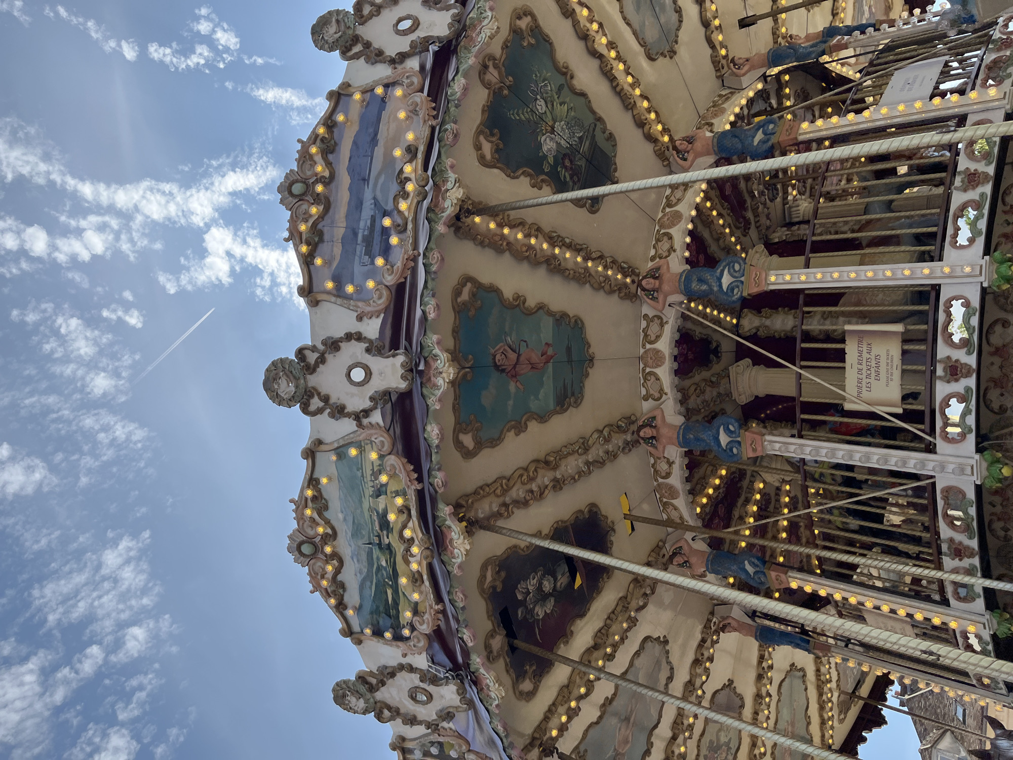 This is a low-angle, close-up shot of a carousel in Rambouillet, France, against a bright blue sky with scattered clouds and a contrail. The carousel is elaborately decorated with painted murals depicting pastoral landscapes, ships, and cherubs. Along the edge of the carousel's roof, a string of small yellow lights is illuminated. Supporting the structure are ornate columns topped with figures of mermaids with blue, scaly tails and human torsos. A sign with French text is visible on a railing within the carousel, partially obscured. The image focuses on the upper portion of the carousel's structure, revealing its intricate details and festive lighting.