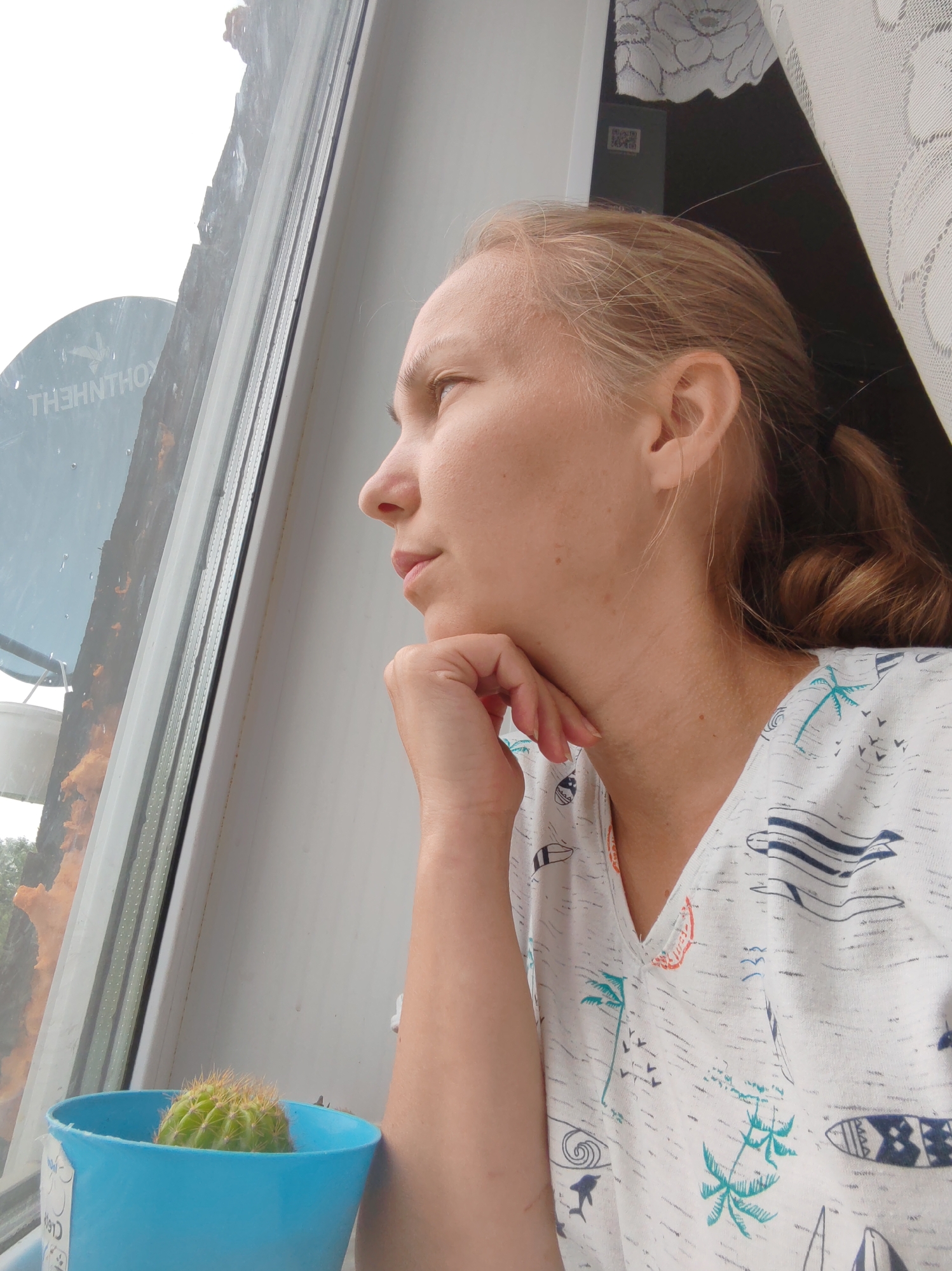 The image shows a woman with light brown hair, looking out a window. She is wearing a white t-shirt with a print of palm trees, surfboards, and birds. Her right hand is resting on her chin. To her left, on the windowsill, is a small green cactus in a blue pot. Outside the window, a satellite dish is visible. The environment appears to be a residential building, possibly in the city of Chukhlem, Russia, given the context. The lighting suggests it is daytime, possibly overcast or rainy, as the window pane has some moisture on it.