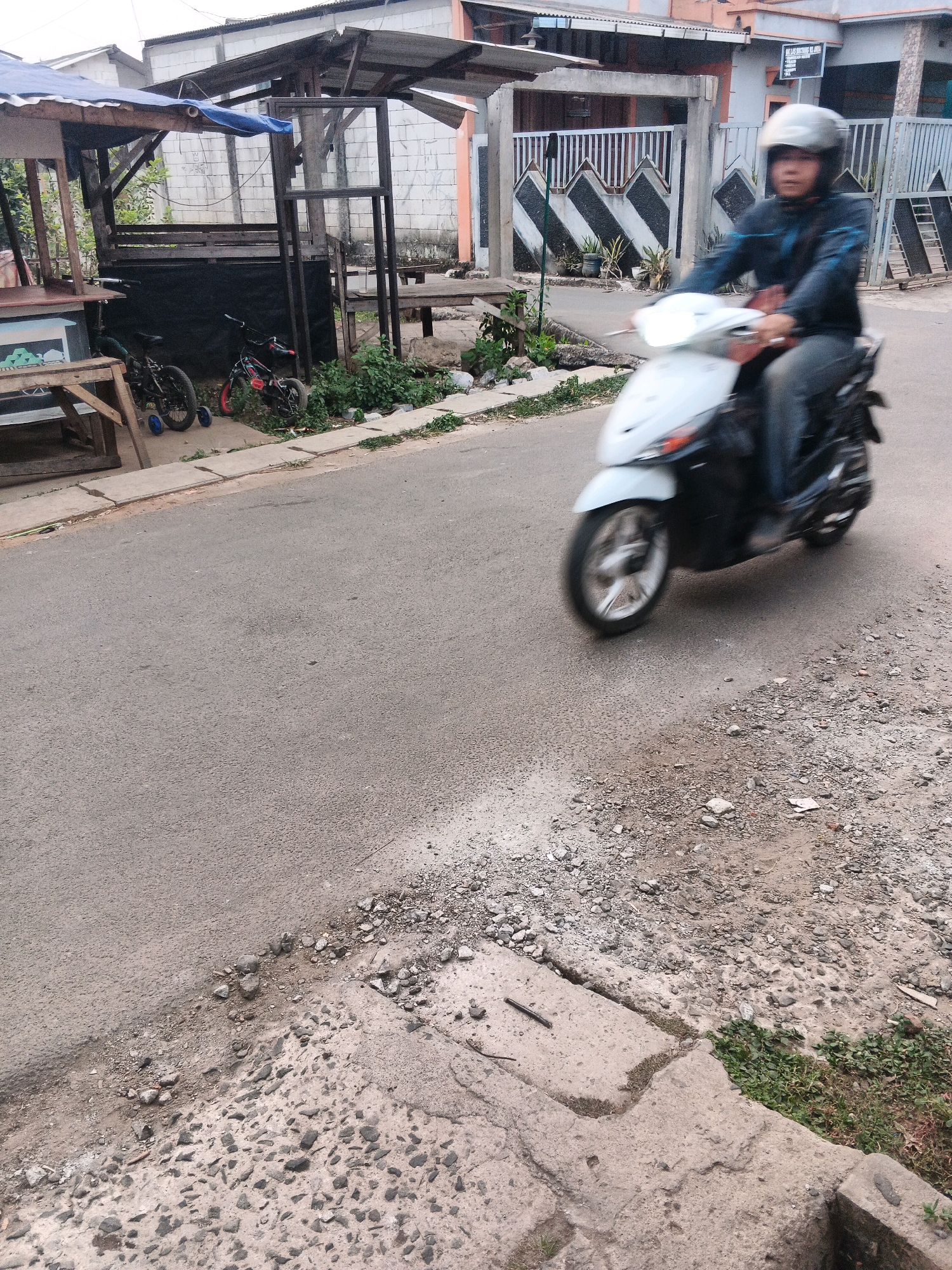 The image depicts a street scene in Kadu, Indonesia. A person wearing a helmet and a blue jacket is riding a white scooter from right to left, with motion blur indicating movement. The scooter has a black lower body and front panel. To the left of the street, there is a small wooden stall with a blue tarp overhead, and two children's bicycles parked nearby. The ground is a mix of asphalt and rougher, rocky terrain with some patches of green vegetation. In the background, a building with a white facade and a grey tiled roof is visible, along with a fence and some potted plants. The overall impression is a casual street environment.