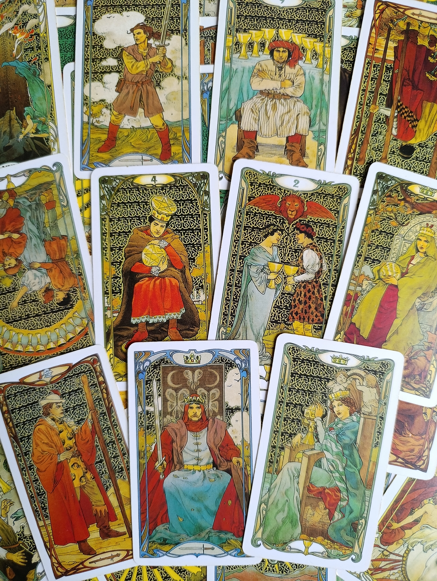 The image shows a collection of tarot cards, laid out in a fanned arrangement against a brightly lit, textured background that appears to be a table surface. The cards feature elaborate artwork in a style that suggests Art Nouveau or a similar decorative period. Each card depicts figures in historical or fantastical attire, often with symbolic imagery.
Several cards are clearly visible and identifiable. One card shows a man holding a sword, standing in a dynamic pose. Another displays a seated figure with a red turban, arms crossed, in front of a row of cups. A card with the number '4' depicts a person in regal attire holding a golden orb. The '2 of Cups' card features two figures offering each other chalices, with a lion-like creature above. Other cards show a figure seated on a throne holding a globe, another figure with a staff, and a queen on a throne. The cards are predominantly colored in earthy tones, reds, greens, and golds, with intricate black and gold decorative borders.
The image does not contain any discernible elements that would suggest a specific location within the city of Alexandrov, Russia, or any other city. The environment is confined to the presentation of the tarot cards themselves.