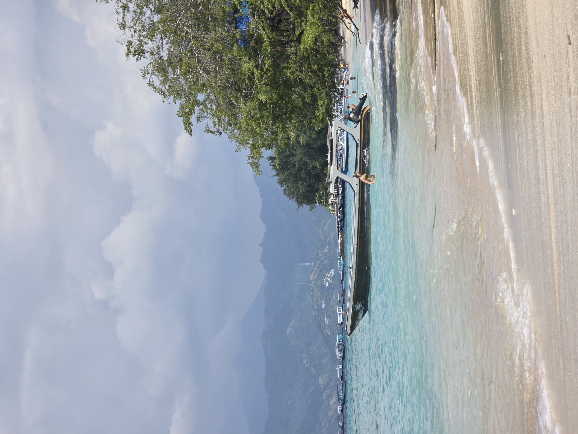 The image depicts a beach scene in Unknown, Indonesia. The scene includes a beach with light-colored sand meeting turquoise water. Several boats are visible in the water, one of which is docked close to the shore with people onboard. In the background, there is dense green vegetation covering the hillside and a cloudy sky above. The waves are gentle, and the overall atmosphere is peaceful and scenic.
