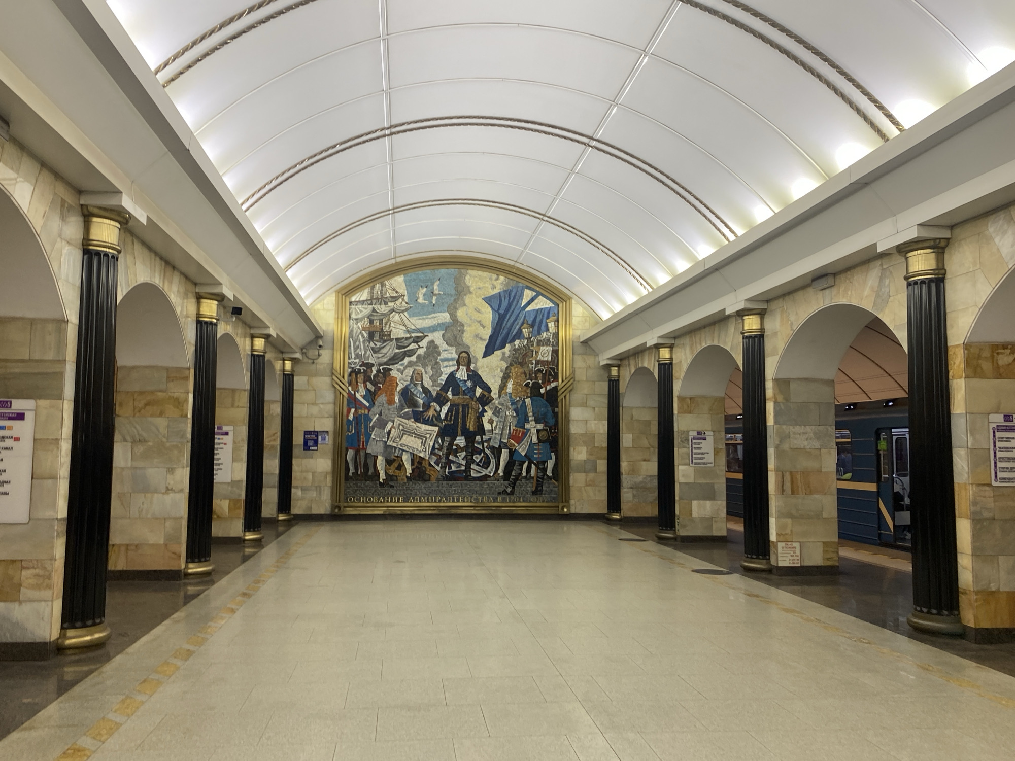 The image depicts the interior of a subway station in Shushary, Russia. A grand mosaic artwork adorns the central wall, featuring historical figures in ornate attire, a sailing ship, and a blue flag. The station's architecture is characterized by a high, arched ceiling with integrated lighting and a series of dark, fluted columns topped with gold accents, separated by arches clad in marble. The floor is tiled in a light color with a decorative yellow strip. To the right, a blue and yellow subway train is visible at the platform, with its doors open. The overall impression is one of spaciousness and historical grandeur.