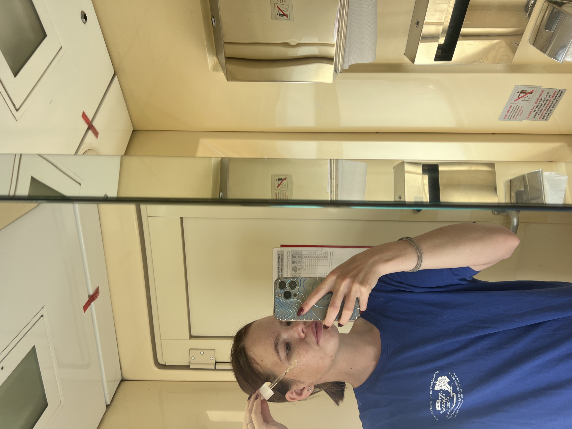 A young woman with freckles and a blue t-shirt is taking a selfie in a mirror inside what appears to be a train compartment. She is holding a dropper with liquid to her cheek, as if applying skincare. Her hair is pulled back in a ponytail, and she is wearing a bracelet and earrings. In the background, a mirror reflects the interior of the compartment, showing a dispenser with paper towels and a sign with a pictogram indicating "no smoking." The walls are a light yellow color, and the ceiling is white with a square panel. There are two red pieces of tape on the ceiling, each forming an 'X' shape. The environment suggests a small, enclosed space, consistent with a train lavatory or a small private compartment. The specific location in the city of Kanash, Russia, cannot be determined from the provided image.
