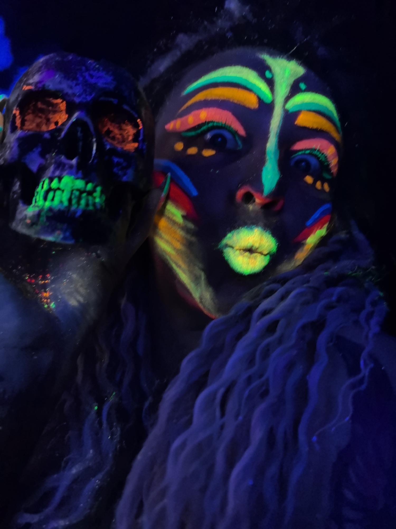 A person with neon makeup stands beside a skull in a dark environment, possibly a Halloween or costume event. The person has colorful neon makeup on their face with intricate designs, including lines and dots. The skull features orange eye sockets and a green mouth. The surroundings are dimly lit, emphasizing the neon colors. Long, wavy hair drapes around the person's shoulders. The image does not reveal information about the location in Noviy, Russia.