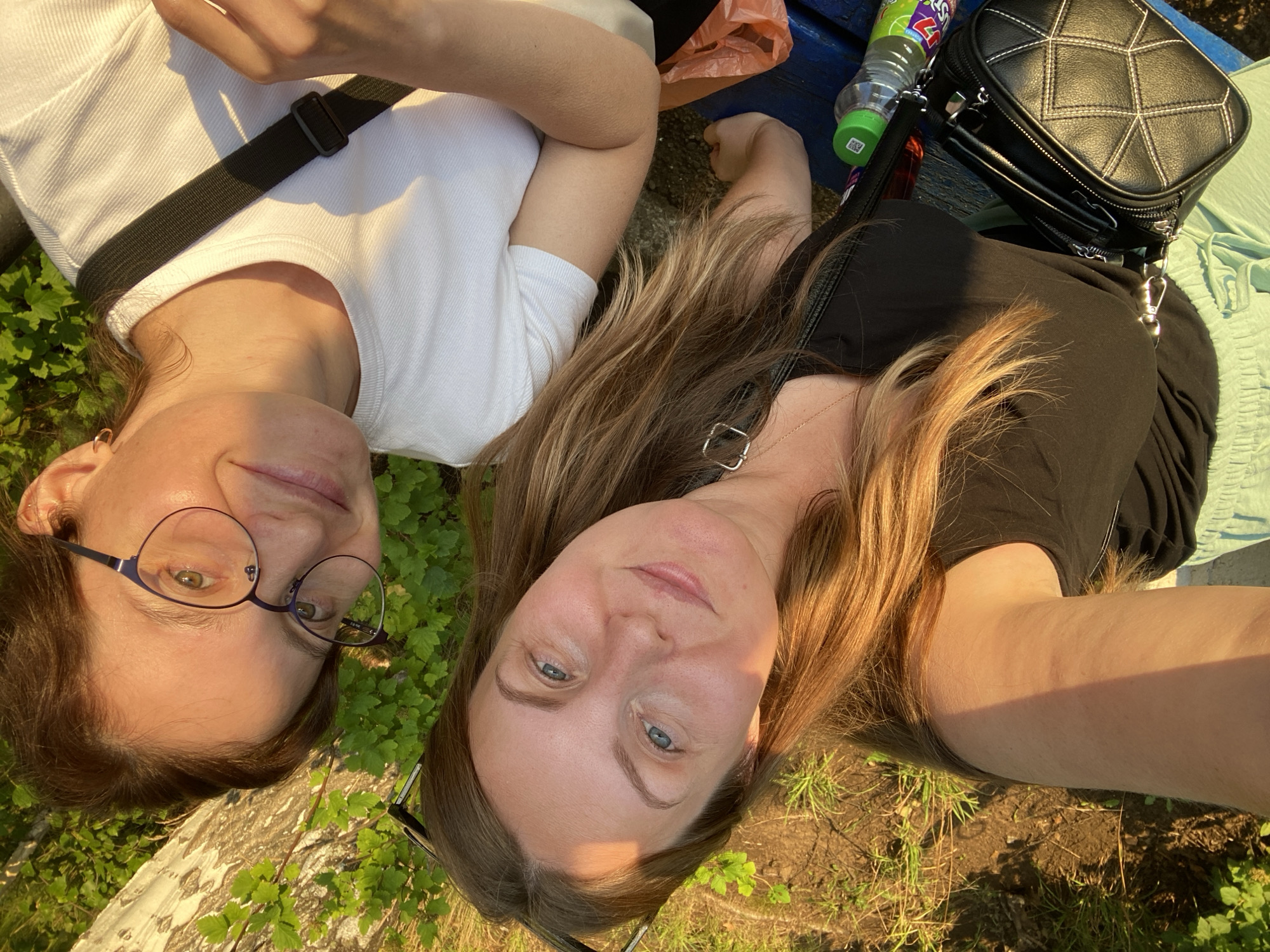 Two women are taking a selfie outdoors in Chaikovsky, Russia. The woman on the left has blonde hair and is wearing a black top. She is holding the camera. The woman on the right has brown hair and is wearing glasses and a white t-shirt. She is smiling at the camera. They are sitting on a blue bench. In the background, there is green foliage and a tree trunk. A black purse, a bottle of juice, and an orange plastic bag are visible on the bench. The lighting suggests it is late afternoon or early evening.