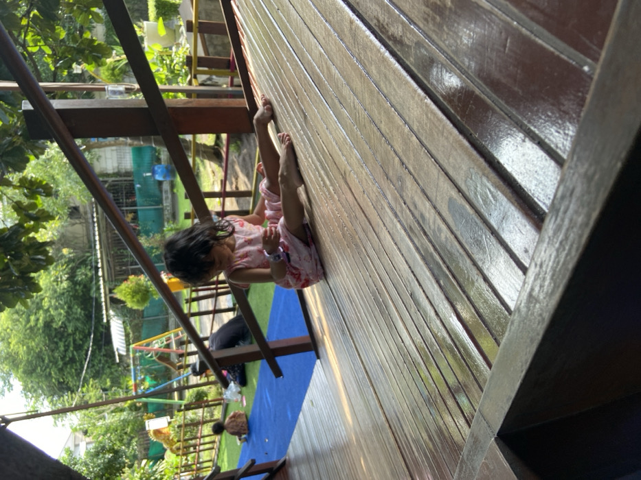 The image shows a young girl in a pink dress sitting on a wooden platform. She is looking down at something in her hand, her feet are outstretched on the platform. The girl is sitting on the platform at the bottom of a wooden ramp. The ramp is surrounded by a wooden railing. The platform is made of wooden planks that are arranged in a grid pattern. The platform is wet and reflects the surrounding area. The image is taken from a low angle, looking up at the girl. The background shows a playground with a swing set and other playground equipment. The scene is located in Kecamatan Sidoarjo, Indonesia (latitude: -7.441, longitude: 112.705).
