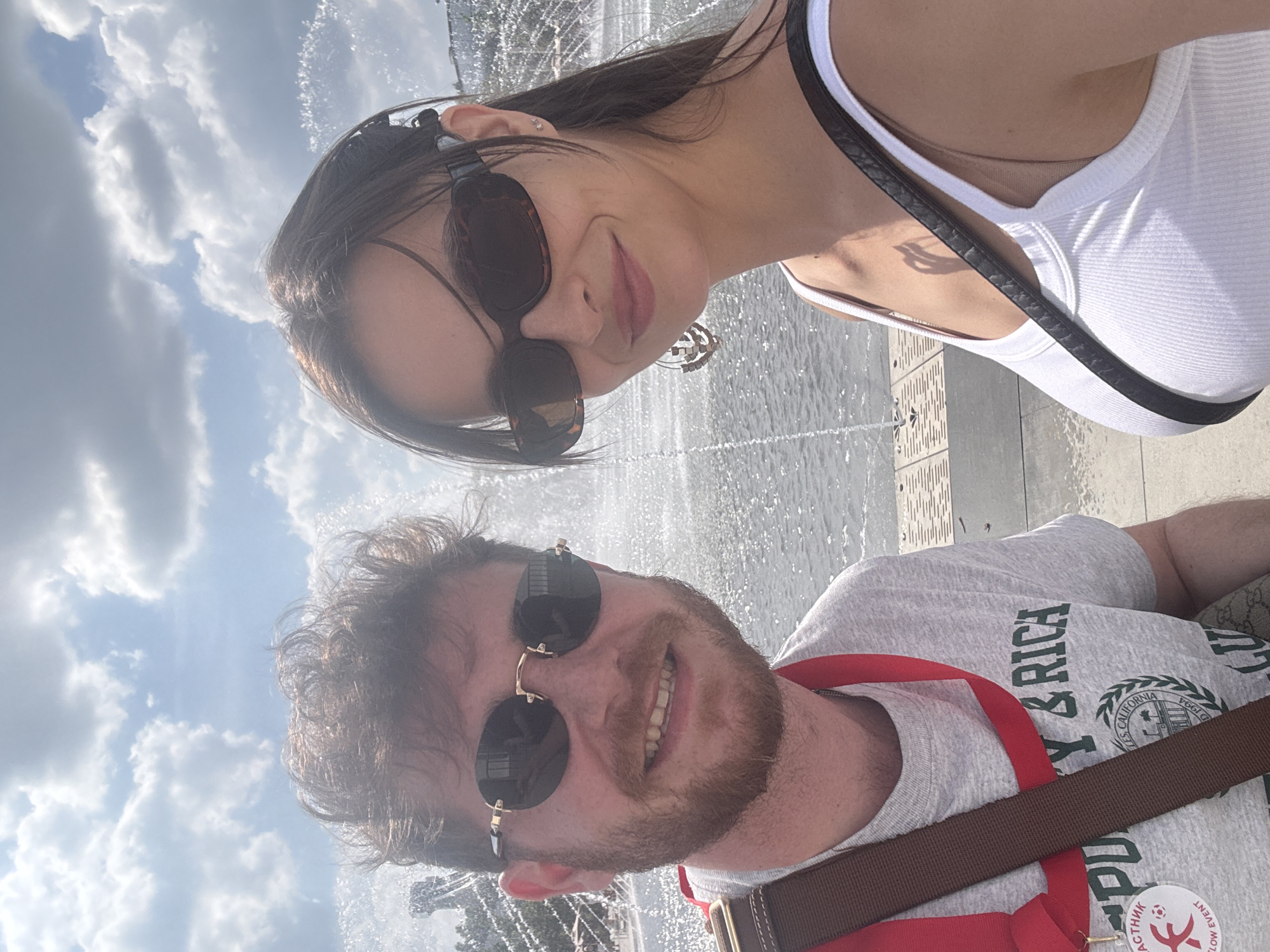 A man and a woman are taking a selfie. The woman is wearing a white tank top and sunglasses. The man is wearing a gray t-shirt with a red lanyard and a brown shoulder bag. They are in an outdoor area with fountains and a cloudy sky. The t-shirt has green text that says "Berkeley & Rica" along with a drawing of the University of California seal, and the medal says "Moscow event." Based on these elements, it seems to be an outdoor area in Moscow, Russia, possibly near a tourist attraction.