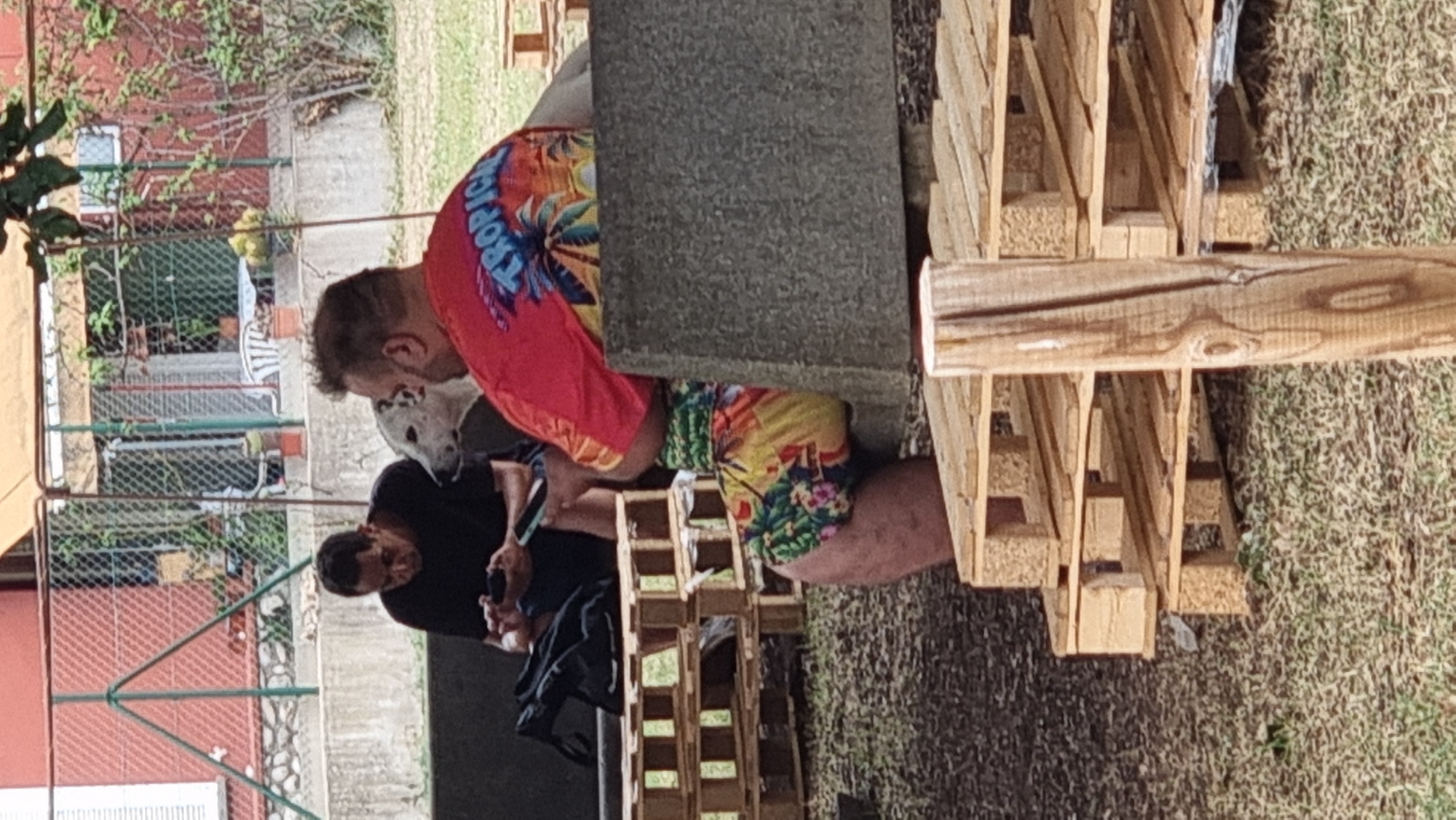 The image depicts two individuals, a man and a woman, seated outdoors. The man, wearing a black t-shirt, is focused on his mobile phone. Beside him, a white dog with black spots rests its head on his lap. The woman, to the right of the man, is wearing a bright red t-shirt with a "Tropical" design featuring palm trees, and colorful floral shorts. She appears to be looking away from the camera. The scene is set in a rustic outdoor environment, with stacks of wooden pallets in the foreground and background. Behind the individuals, a chain-link fence encloses a yard with a building and some greenery. The ground is covered with grass and dry leaves. The lighting suggests it is daytime, possibly overcast. The overall atmosphere appears casual and relaxed.