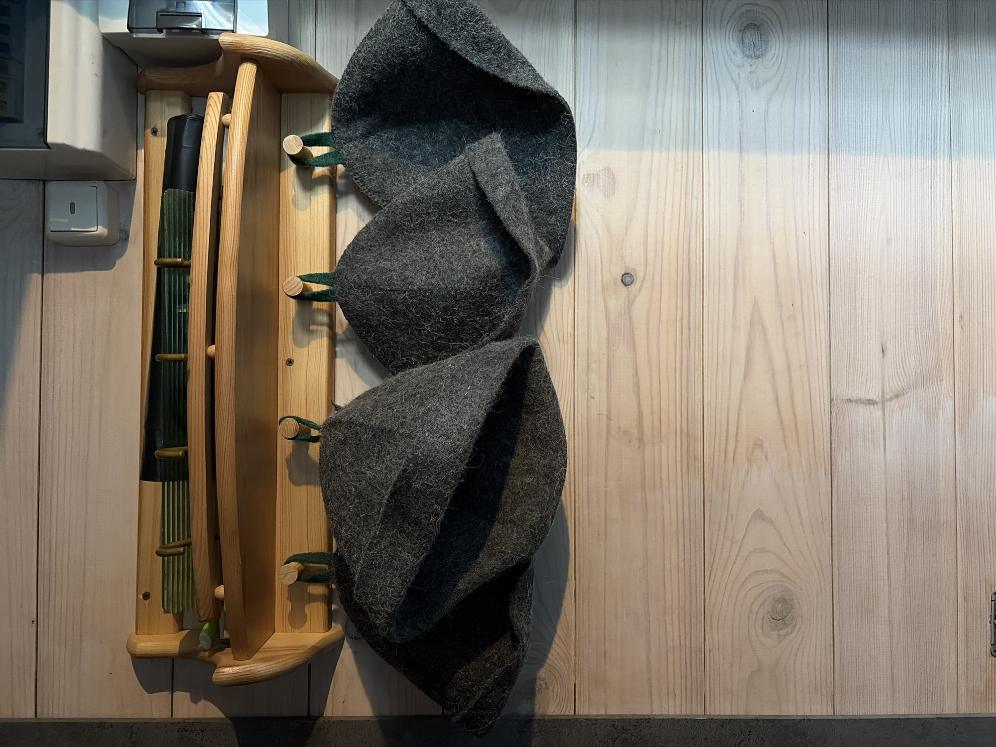 A wooden shelf with a rounded front is mounted on a light-colored wooden wall. On the shelf are two items: a black cylindrical object with green fins and a black tubular object with some green markings. Below the shelf, four dark gray felt hats with green loops hang from wooden pegs. The hats are shaped like inverted buckets. The wall appears to be made of horizontal planks.