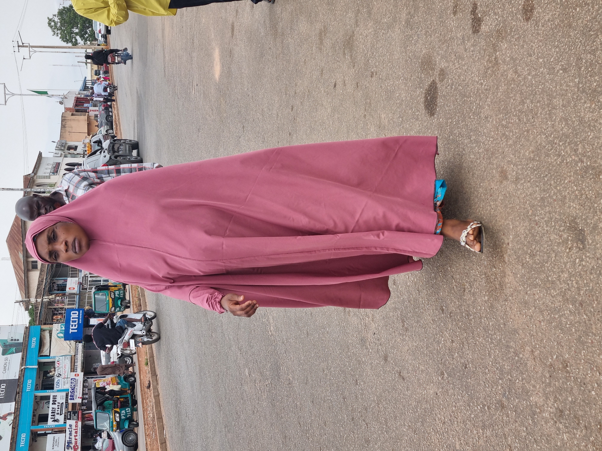 A vertical full-body shot captures a woman in a bustling street scene in Jos, Nigeria. She stands slightly to the left of the frame, facing the camera with a neutral expression. She wears a long, flowing pink abaya or hijab that covers her head and body, reaching her ankles, with a lighter blue pattern visible underneath at the bottom edge. White sandals are on her feet. Her right hand is slightly open, palm facing upwards.
Behind her, an older man in a plaid shirt is partially visible. The background reveals a lively street lined with single and multi-story buildings, characteristic of a commercial area or market. Numerous vehicles, including motorcycles (often referred to as "okadas" or "bajaj" tricycles, locally known as "keke napeps," with "PTA 9113" and "PTA 6436" seen on two of them) and cars, are parked or moving along the road. Several people are visible in the background, some walking, others on motorcycles. The ground is a light-colored asphalt.
The time of day appears to be daytime, with bright ambient light suggesting either overcast conditions or a bright, non-midday sun, as shadows are not prominent. A Nigerian flag is faintly visible in the upper left background.
Visible text on the buildings and signs includes multiple instances of "TECNO" (likely a phone brand), "CAMON 50i," "KINSHANG GLOBAL," "EARLY DOVE," "STERE," "DSCCTV GOL," "BRISKHUO PHONES LAPTOPS ACCESSORIES," "GREG ELECTRONICS," and "Miracle Curtains."