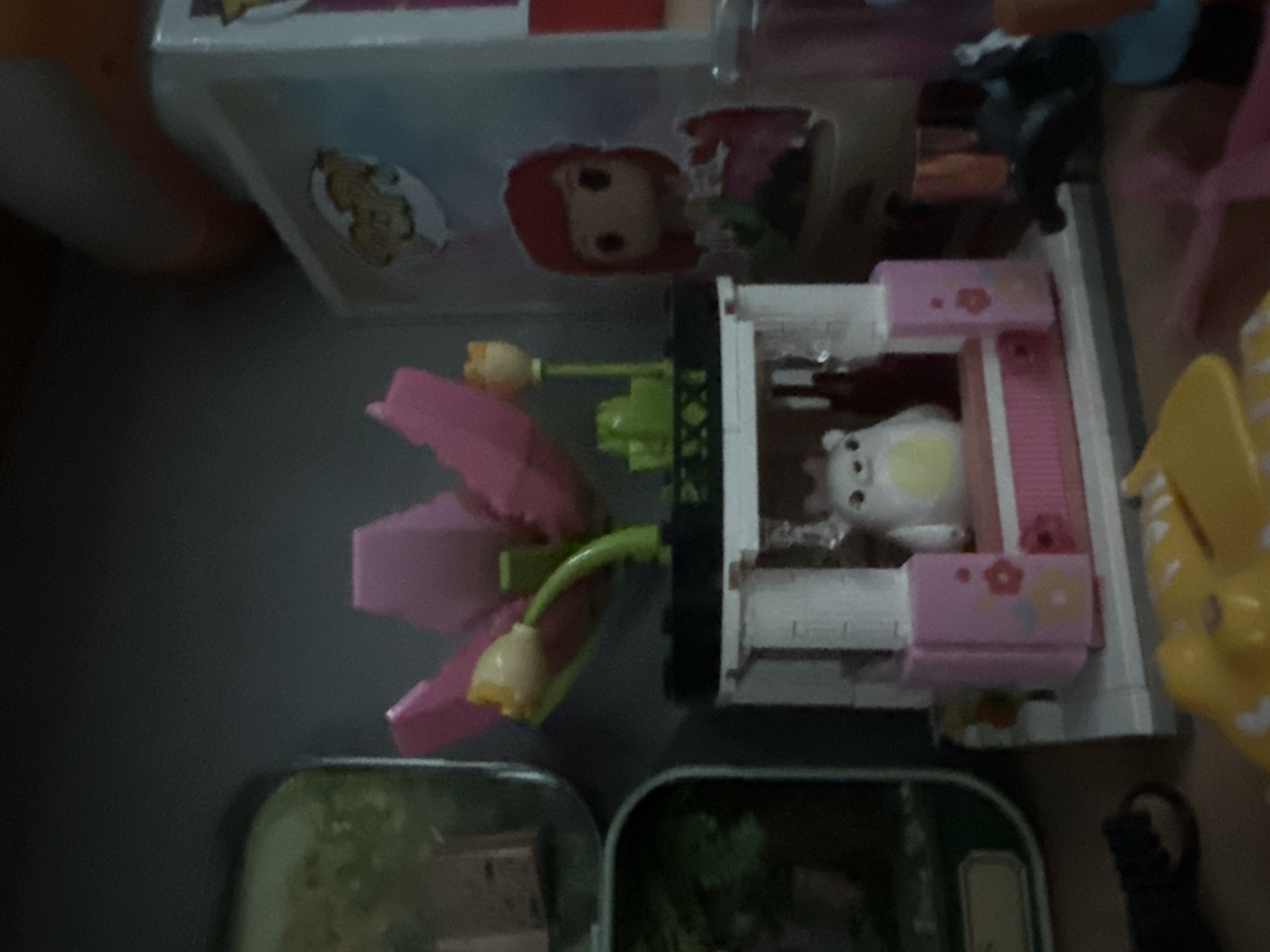 The image shows a collection of toys, including a Lego flower stand with a small white bear figure. There is also a Funko Pop figure of a character with red hair and a pink dress, and what appears to be other Lego structures and figurines. The background is dark, suggesting indoor lighting. The image does not contain any information that would allow for the determination of its location within the city of Petropavlovsk-Kamchatsky, Russia.