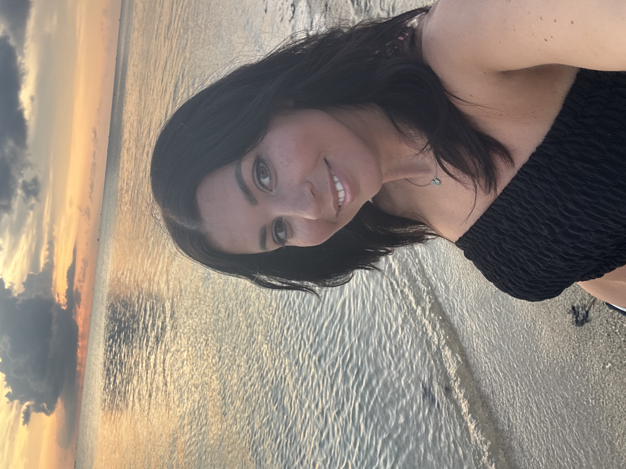 A woman with dark hair smiles at the camera. She is wearing a black strapless top. The location appears to be a beach at sunset in Baan Sri Thanu, Thailand. The water reflects the golden light of the setting sun. The sky contains both orange and grey clouds. Sand is visible at the water's edge.