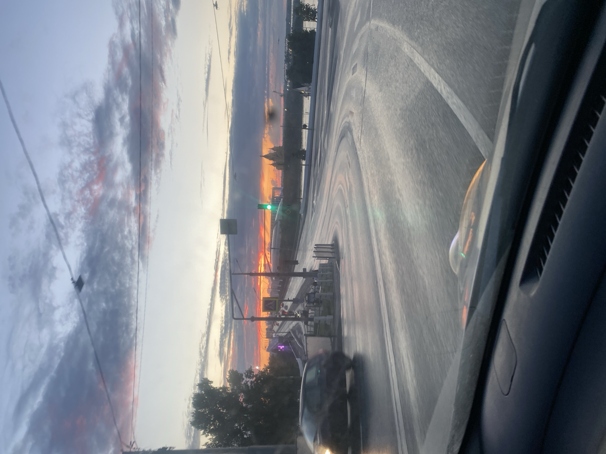 This is a vertical, handheld, point-of-view photograph taken from inside a vehicle. The image depicts a sunset over a road and distant cityscape. The sky is filled with dramatic clouds, tinged with orange, pink, and gray, indicating it is either sunrise or sunset. A traffic light showing green is visible on the left side of the road.
In the foreground, the interior of the vehicle is partially visible, including a dark dashboard and a window. The road ahead is wet, reflecting the sky and the lights from the setting sun. A car is in motion on the road, blurred due to its movement. The distant horizon shows the silhouettes of buildings, including what appears to be a prominent church or cathedral with multiple domes, characteristic of Russian architecture. Trees are visible on the left side of the road, silhouetted against the bright sky. The overall mood of the image is serene and atmospheric, capturing the beauty of a sunset in an urban or semi-urban environment. The location is confirmed as Nizhny Novgorod, Russia.
