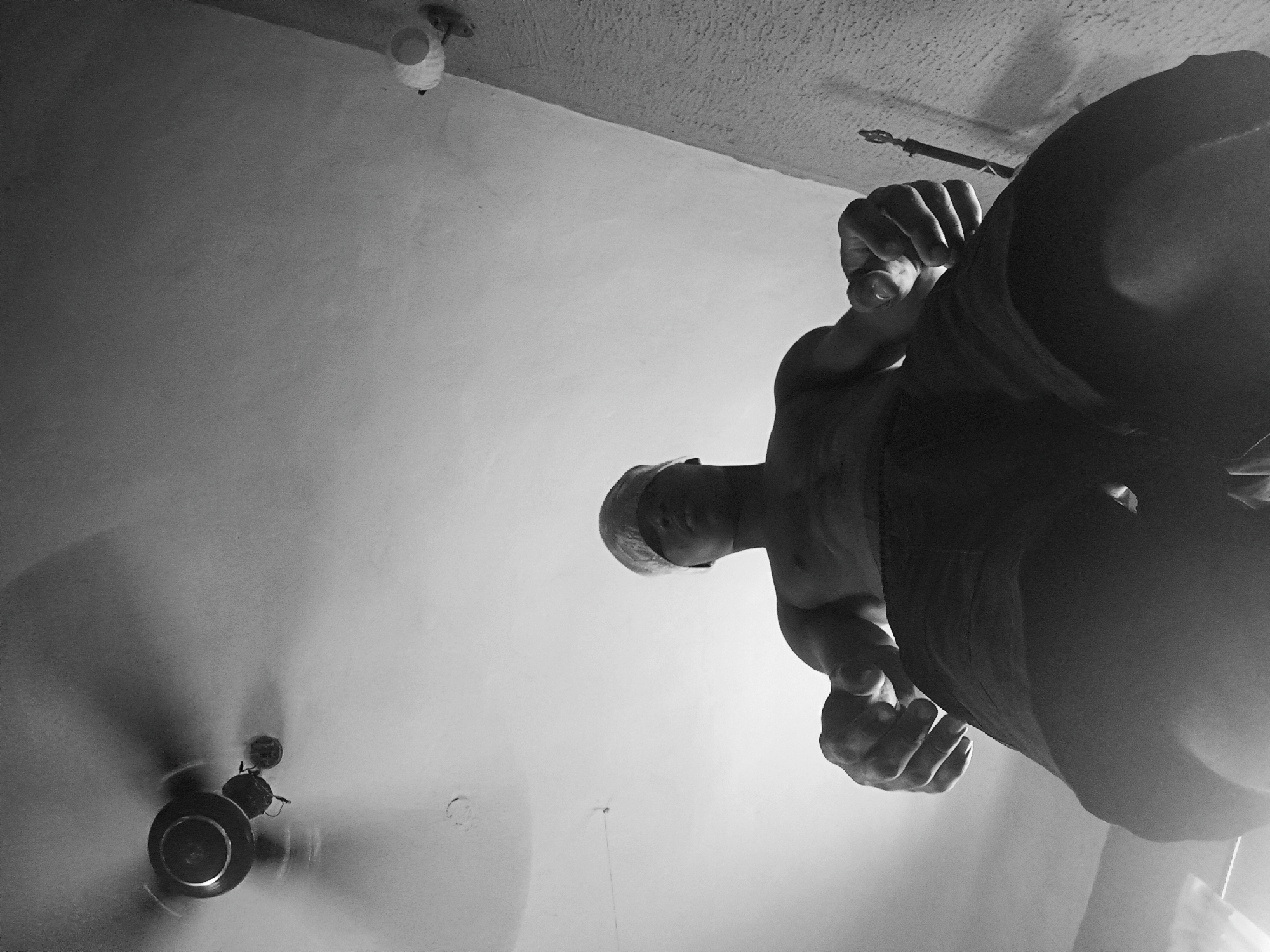 A black and white, high-angle photograph shows a man looking down at the camera, seemingly from a low perspective. The man has a shaved head and is wearing dark clothing. His hands are visible near the bottom of the frame. Above and to the left, there is a ceiling fan with blurry blades. Also visible on the ceiling is a light fixture. Nkpor, Nigeria is not indicated in this image.