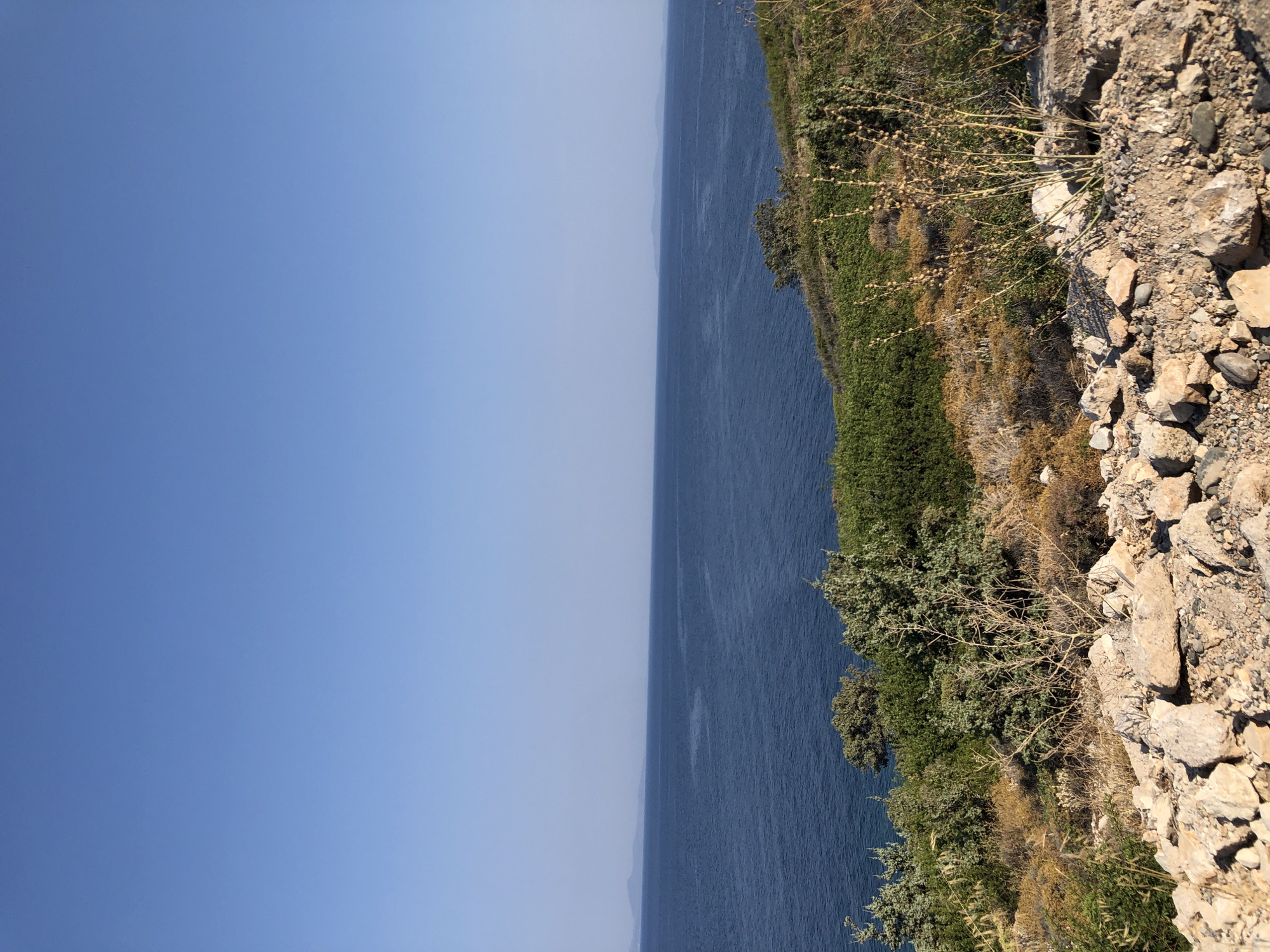 The image shows a wide view of the sea and coastline. In the foreground, there are rocks and dry vegetation on a hillside. Behind this, there are green bushes and trees. The sea is a deep blue, and in the distance, there are faint outlines of islands or land. The sky is clear and blue. The scene appears to be in a natural, coastal environment. The location is identified as Karaburun, Turkey. There are no people or specific actions depicted in the image.
