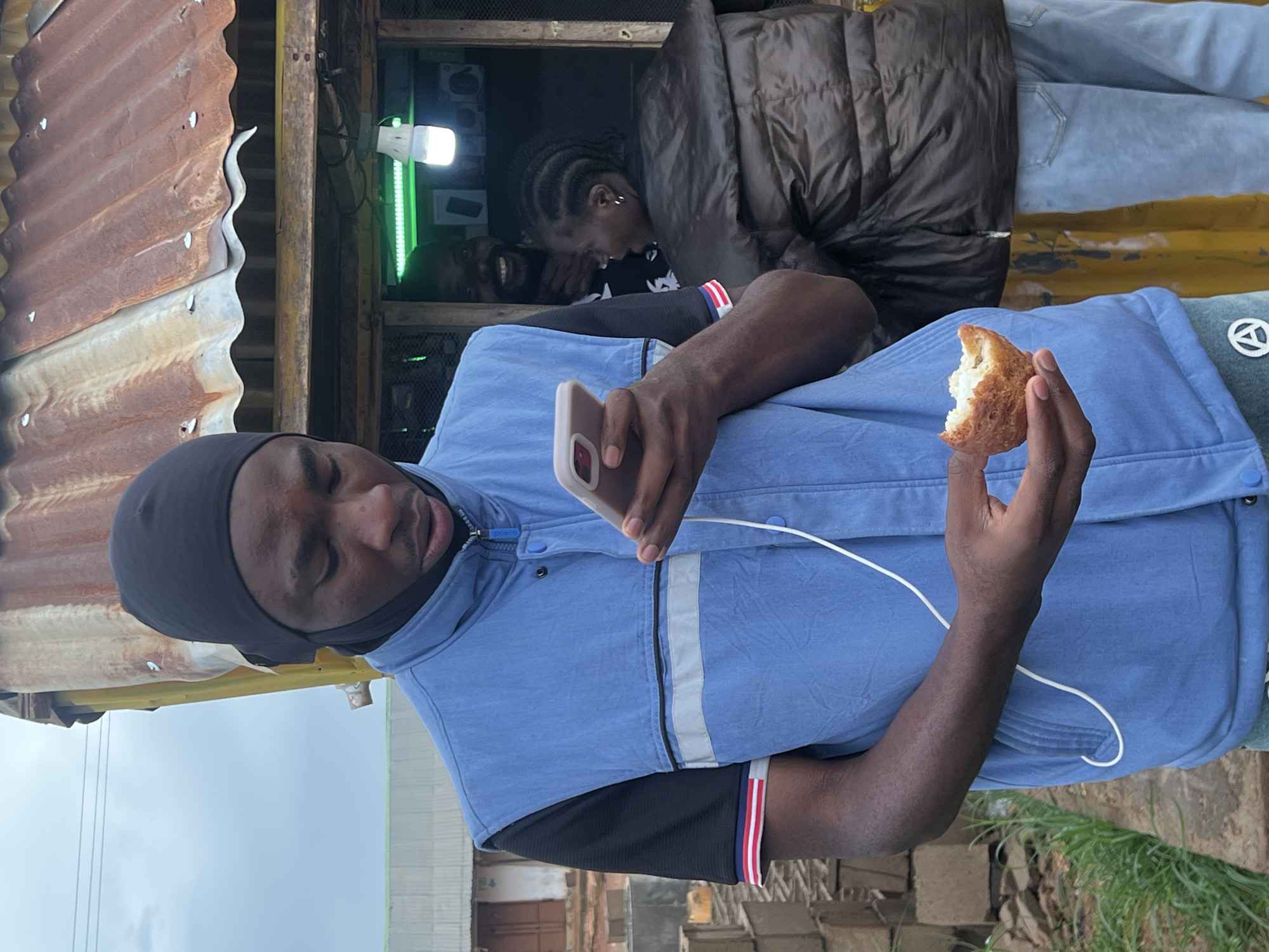 A young Black male wearing a blue vest over a black t-shirt and a black head covering looks down at his phone while holding a piece of food in his right hand. He is standing in front of a small roadside kiosk with a corrugated metal roof. Behind him, a Black man with a beard is smiling and looking at the camera, and a Black woman in a dark puffer jacket and light blue jeans stands next to him. The kiosk appears to be selling electronics, as indicated by a bright green light and several items displayed inside. The background shows some construction materials and a building. The overall scene suggests a casual outdoor setting in a Nigerian city.