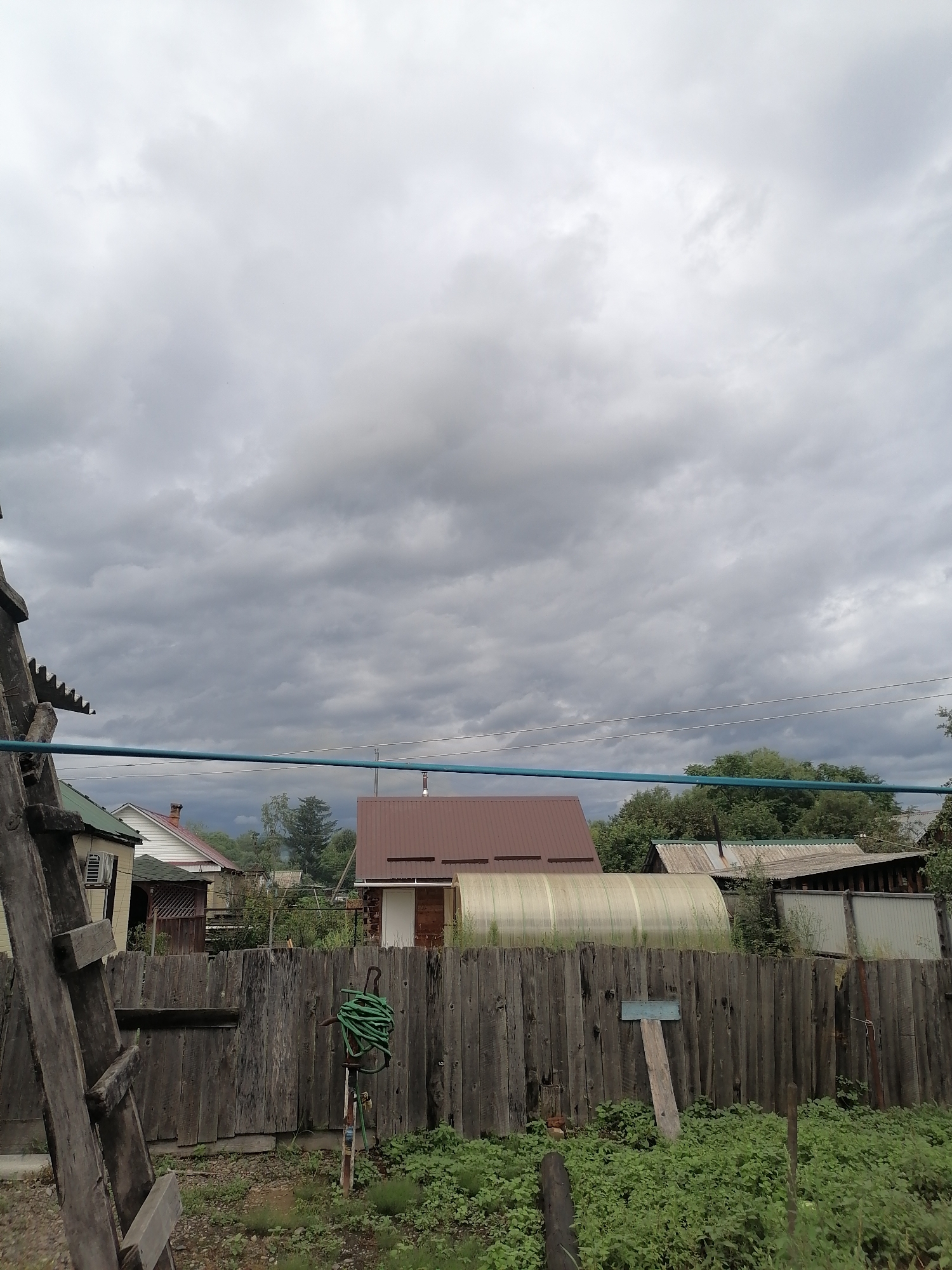 The image depicts a cloudy sky over a rural village setting, likely in Novopokrovka, Russia. In the foreground, a weathered wooden fence runs horizontally across the frame. To the left, a portion of a wooden ladder is visible, leaning against an unseen structure. A green garden hose is coiled around a vertical pipe affixed to the fence. Beyond the fence, several structures are visible, including a house with a green roof and white siding, a greenhouse with a curved, translucent roof, and a building with a brown, gabled roof. Trees and vegetation surround the buildings, and electrical wires are strung across the sky in the upper right quadrant. The overall scene suggests a somewhat overcast day in a residential area.