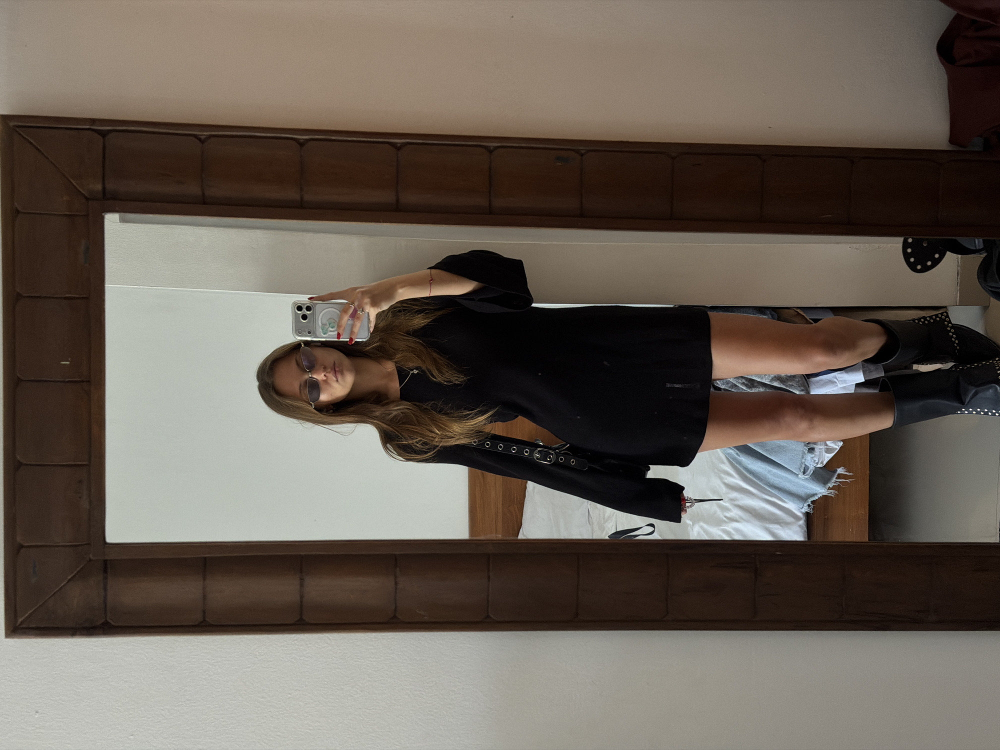 A woman is taking a selfie in a mirror. She is wearing a black dress and boots, and has sunglasses on. The mirror has a wooden frame. The location appears to be a bedroom. There is a bed behind her. There is no confirmed information about the location being in North Kuta, Indonesia.