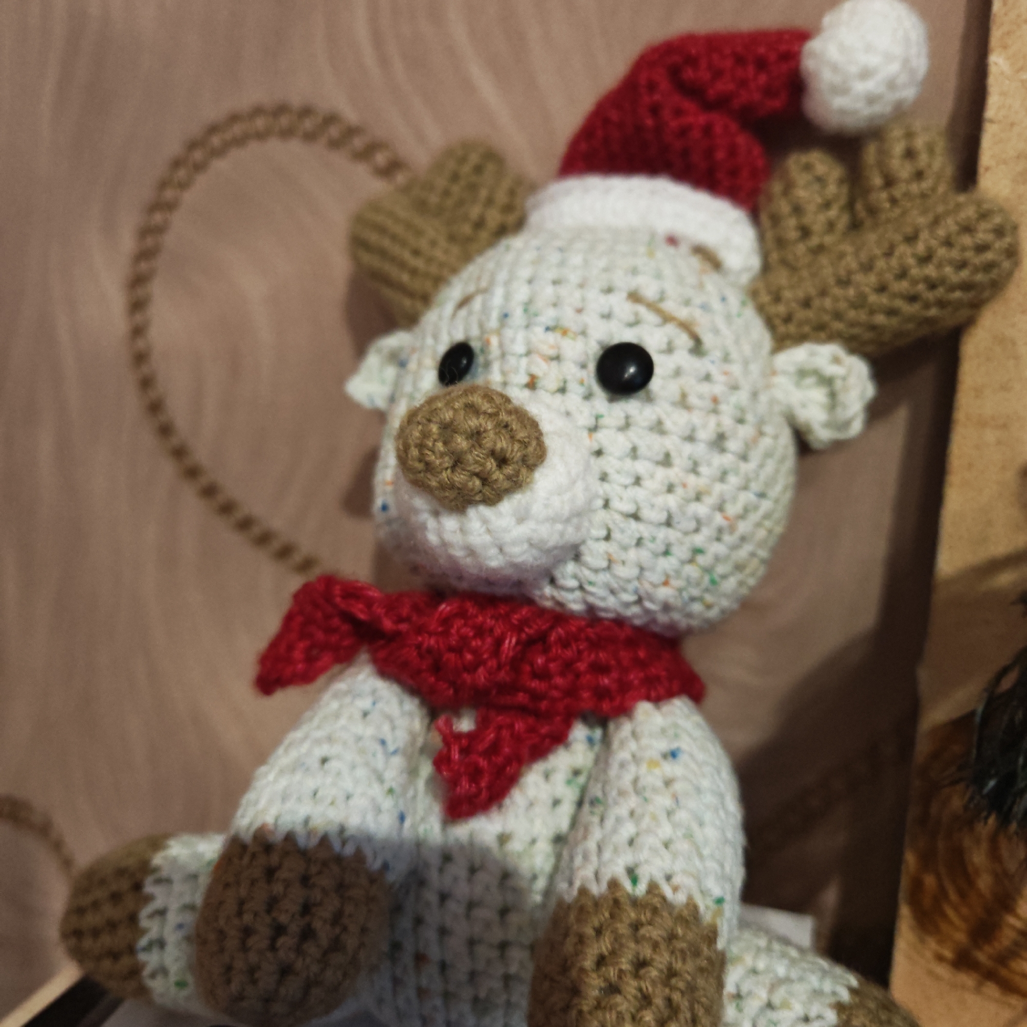 A close-up shows a crocheted reindeer wearing a red Santa hat with a white pompom, a red scarf, and brown hooves and antlers. The reindeer has large, black, button eyes and a brown nose. The body is cream-colored with flecks of color in the yarn. In the background, a wooden surface with a rope design is visible. The location is Rostov, Russia.