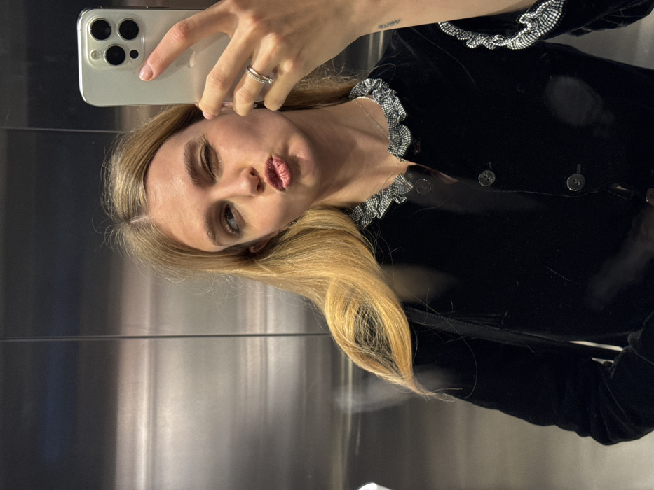 A blonde woman takes a selfie in an elevator with her iPhone. She is wearing a black velvet jacket with a ruffled collar and buttons. Her lips are pursed, and one eye is closed. She wears a silver ring on her finger. The elevator walls are mirrored. A small tattoo is visible on her arm.