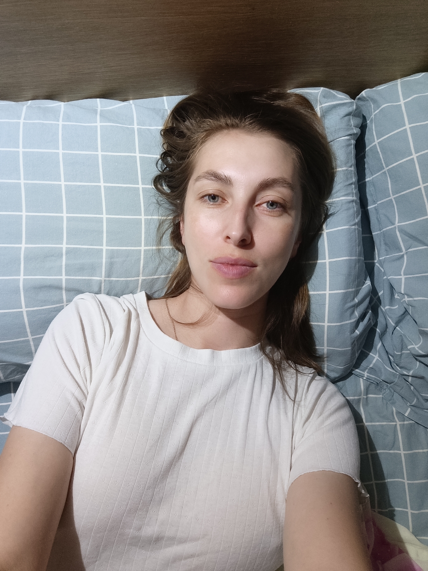 A young woman with light brown hair and blue eyes is lying on a bed with blue and white checkered bedding. She is wearing a white t-shirt. The headboard of the bed is visible at the top of the image. The scene is indoors, likely a bedroom. The lighting suggests it is nighttime or a dimly lit room. There is no indication of the city of Chaikovsky, Russia, in the image itself.
