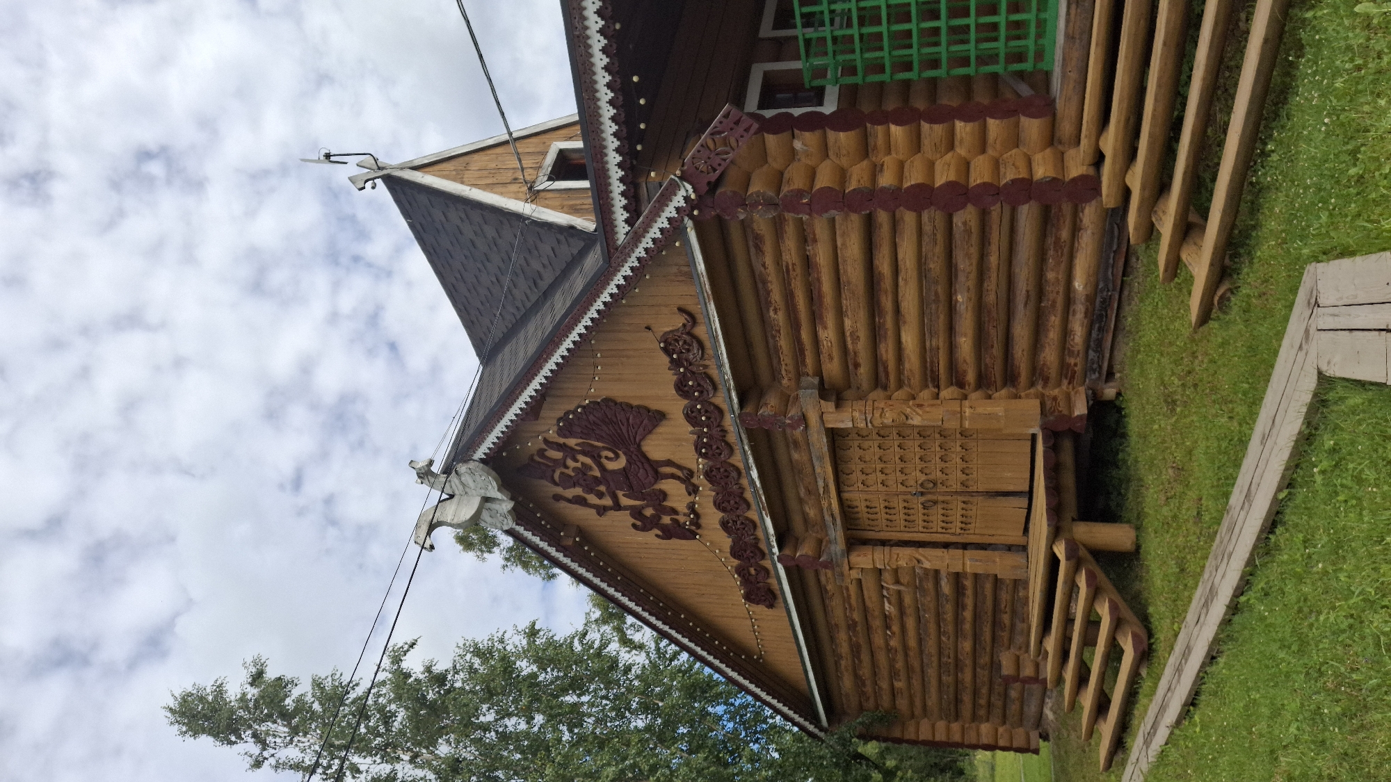 The image shows the side of a traditional wooden house with intricate carvings. The house is constructed from round logs and features decorative wooden elements under the eaves. One carving depicts a stylized bird, possibly a phoenix or a rooster, in a deep red color. The roof is dark and shingled, and a pointed gable rises above. A small attic window is visible. A wooden staircase leads up to a small porch or entrance. Part of a green latticework structure is visible at the top of the house. The house is surrounded by a grassy area, with trees visible in the lower left corner. The sky is overcast with clouds. There is no text visible in the image. The scene appears to be outdoors, possibly in a rural or historical setting. The lighting suggests it is daytime, and the cloudy sky indicates the weather is not sunny.