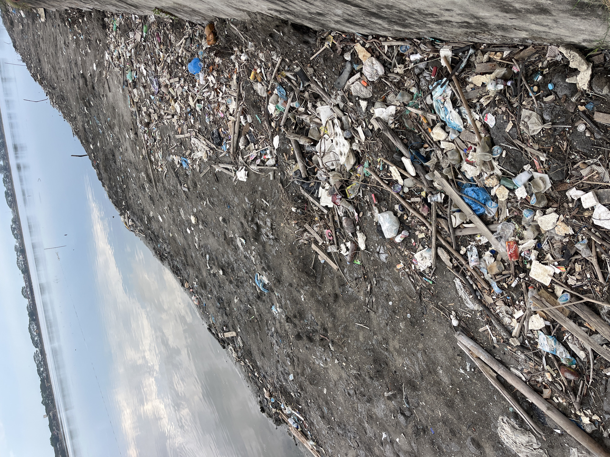 The image shows a heavily polluted area in Lhokseumawe, Indonesia. A body of water is visible to the left. The shoreline and adjacent land are covered in a dense layer of mixed trash, including plastic bottles, bags, styrofoam, wood debris, and other unidentifiable waste. A concrete wall or structure is visible along the top edge of the image. The ground appears muddy and dark. There are no people visible in the scene. The overall impression is one of significant environmental degradation.