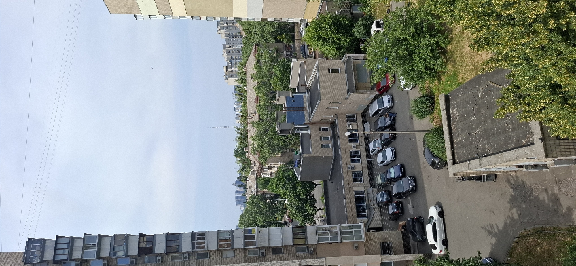 This is an aerial shot of a residential area in Kyiv, Ukraine, featuring a mix of apartment buildings and smaller structures. The foreground is dominated by the side of a tall, multi-story apartment building on the left, characterized by numerous balconies and windows. To the right of this building, a lower, light-colored brick building with a flat roof is visible, with a parking lot in front of it. Several cars are parked in the lot, including a white sedan, a dark-colored sedan, a blue sedan, a silver minivan, and a red car. Further into the background, more apartment buildings and trees fill the landscape, suggesting a densely populated urban environment. The sky is overcast with some faint clouds. A few people are visible on the street and sidewalks. The scene appears to be a typical urban scene during the daytime.