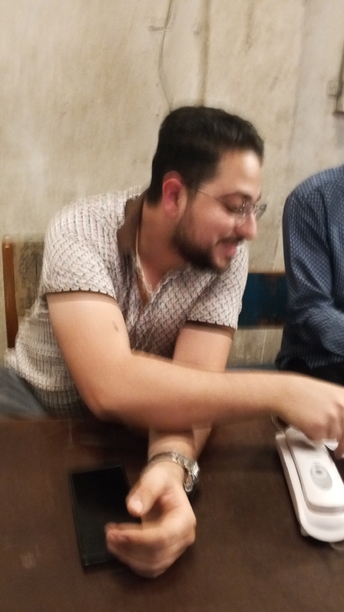 This image depicts an interior scene in the city of Dakata, Nigeria, featuring two individuals seated at a dark brown table. On the left, a male subject with dark hair, a beard, and glasses is seen from the chest up, wearing a light-colored patterned polo shirt with a brown collar. He is smiling or laughing, looking downwards and to the right. His left arm rests on the table, displaying a silver-colored wristwatch. A black smartphone is positioned face down on the table near his left hand. His right arm is extended across the table, with his hand partially obscured. To the right, a second individual is partially visible from the chest up, wearing a dark blue patterned shirt. This person's right hand is interacting with a white electronic device, possibly a landline phone receiver, which rests on the table. The background consists of a plain, light-colored wall with faint texture, and a horizontal blue element is visible behind the second individual. The lighting is dim.