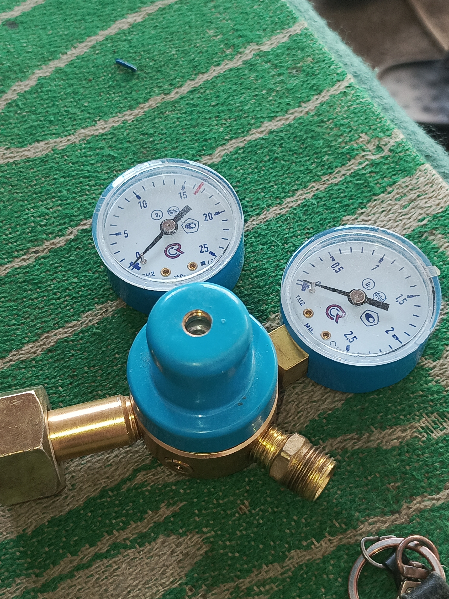 The image shows a close-up, high-angle shot of a gas pressure regulator with two gauges, placed on a green and white striped cloth. The regulator has a blue top and brass fittings. The gauge on the left shows a reading between 20 and 25 units, and the gauge on the right shows a reading between 0.5 and 1.0 units. A small blue object is visible on the cloth in the upper left quadrant of the image. The overall environment appears to be indoors, possibly a workshop or storage area, in Kumkurgan, Uzbekistan. There are no people or actions depicted in the image.