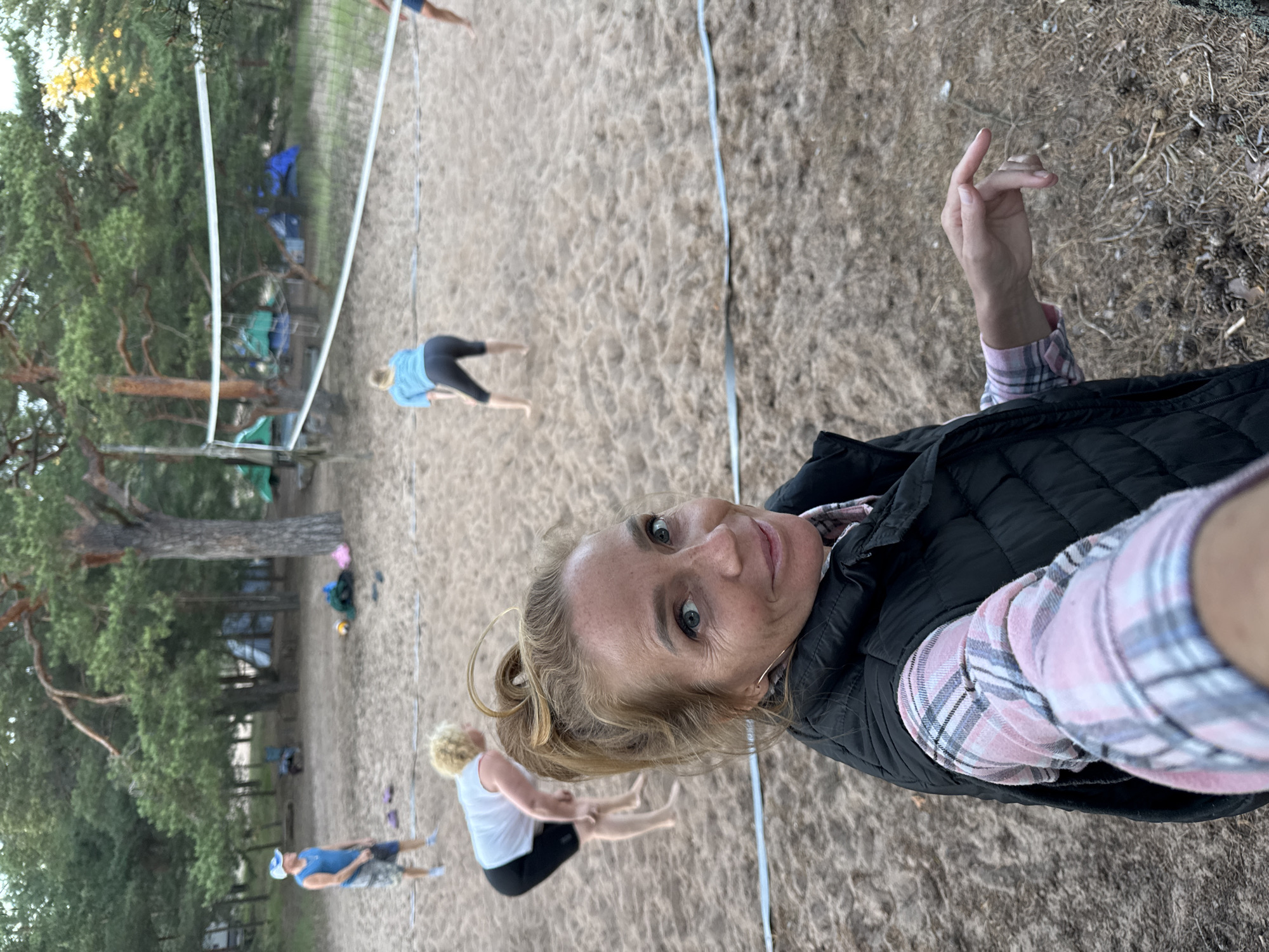 In Ozerki (Krasnodolinskaya), Russia, a group of people are playing volleyball on a sandy court. In the foreground, a woman with blonde hair tied in a ponytail, wearing a pink and white plaid shirt and a black vest, smiles at the camera. She is positioned to the right of the frame, with her left arm extended towards the viewer. In the background, three other individuals are visible. One woman with blonde hair, wearing a white tank top and black shorts, is bent over on the sand. Another woman, also with blonde hair, is in the background, wearing a blue long-sleeved shirt and black leggings, in a volleyball stance. A man wearing a blue tank top and patterned shorts is standing further back on the left side of the court. The court is surrounded by pine trees, and several tents are set up in the background, suggesting a campsite or park setting. A volleyball net stretches across the middle of the court. The ground is covered in sand, with scattered pine needles and cones. The lighting suggests it is late afternoon or early evening.