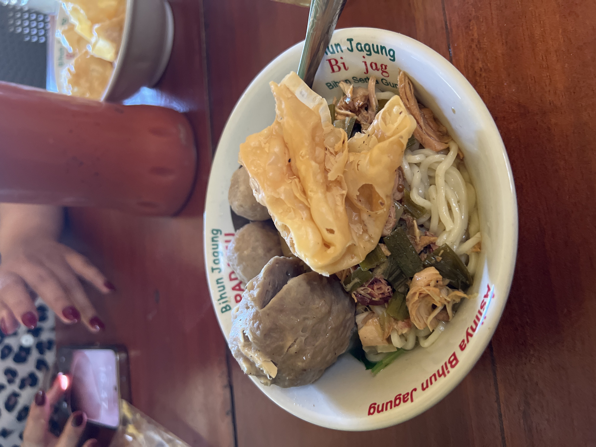 A close-up shot shows a bowl of noodle soup on a wooden table. The bowl has red text printed on its rim. The soup contains meatballs, noodles, and shredded chicken. A fried wonton is perched on top of the noodles. In the background, a person's hands are visible, one holding a smartphone. A bottle of red liquid and a small bowl of snacks are also present. The scene appears to be in Gianyar, Indonesia.