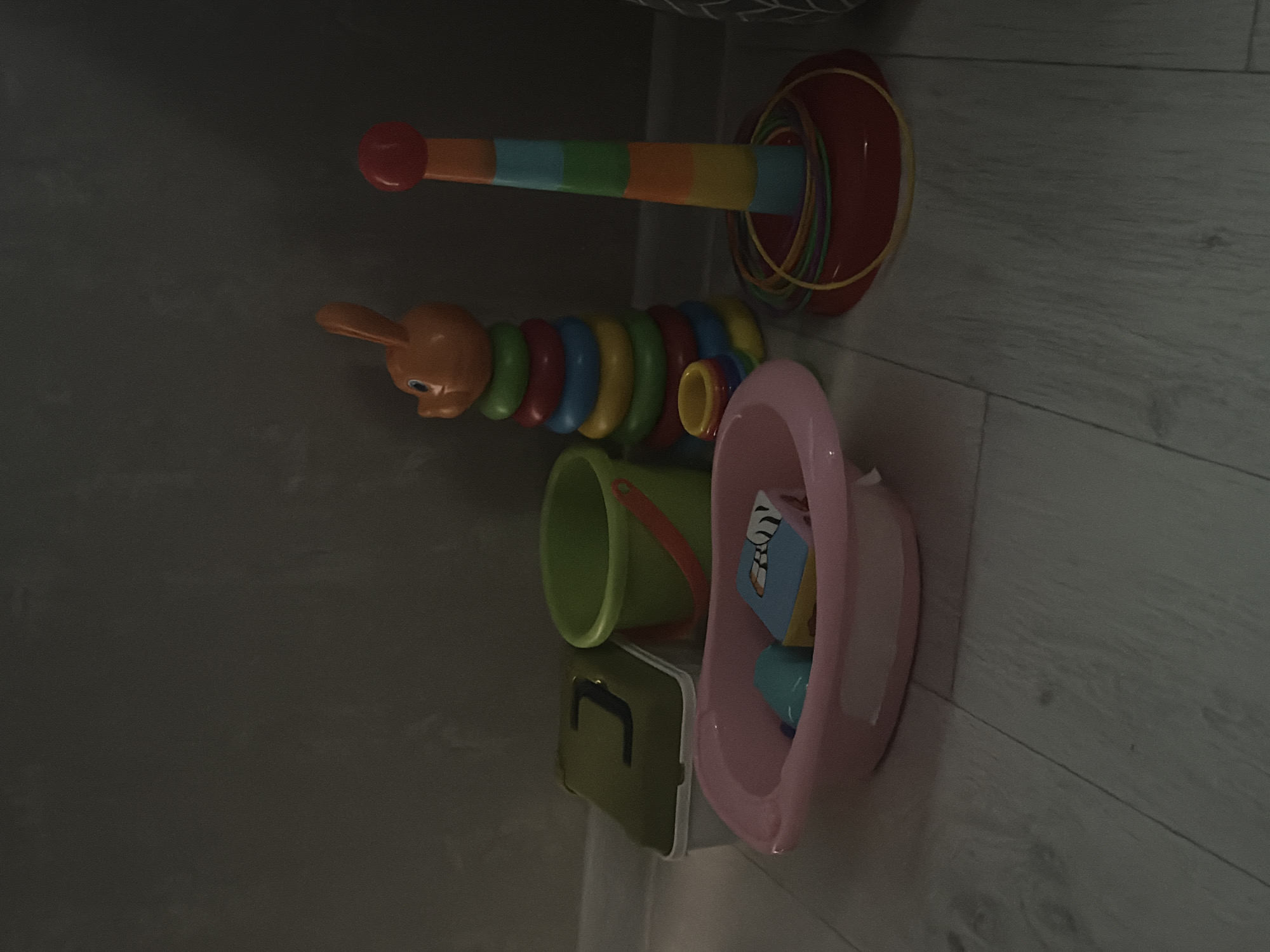 This media file features a collection of children's toys arranged on a light-colored wooden floor. The primary objects are a stacking ring toy with colorful rings and a red base, and a segmented caterpillar toy with an orange head and colorful rings. Next to these, a green bucket with an orange handle and a small green lidded container are visible. A pink bathtub-shaped toy, containing a few smaller colorful objects, sits to the right of the green bucket. The background is a dark, textured wall, possibly a closet or wall surface, which obscures much of the setting. The lighting suggests an indoor environment, likely a room with low ambient light, possibly an evening or a dimly lit area. The overall scene depicts a play area, hinting at the presence of children. No people are visible in the image. There is no visible text in the media file. The location context is Krasnoyarsk, Russia.