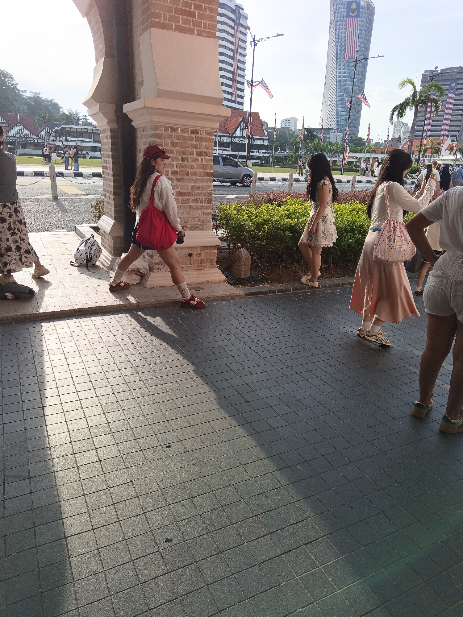The image captures a daytime scene outside the Sultan Abdul Samad Building in Kuala Lumpur, Malaysia. The foreground is dominated by a paved walkway with a tiled pattern, reflecting sunlight and casting distinct shadows. In the midground, several individuals are visible. To the left, a person wearing a patterned skirt and a light-colored top walks away from the camera. Beside them is a backpack with a light-colored pattern on a dark background. Further to the right, a woman with long dark hair and a red cap is seen from the waist down, wearing a white top, dark shorts, white socks, and red shoes, carrying a large red tote bag over her shoulder and leaning on one leg. She appears to be looking to her right. To her right, another woman with long dark hair, wearing a white floral print dress and white shoes, walks to the left. She is partially obscured by a dense green hedge. Beyond the hedge, a woman with long dark hair is seen from the waist down, wearing a white long-sleeved top, a pale pink skirt, and white socks with brown shoes, holding a phone in her right hand and a pink patterned bag over her left shoulder, appearing to take a photograph. Partially visible at the far right edge of the frame is the lower half of another person wearing white shorts and shoes. In the background, the iconic architecture of the Sultan Abdul Samad Building, with its brick facade and arched entrance, is prominent. Beyond that, modern skyscrapers, including the iconic Felda Tower draped with Malaysian flags, rise into a bright blue sky. Other buildings with traditional colonial architecture and Malaysian flags are also visible, along with trees and lampposts. The overall atmosphere suggests a tourist area on a sunny day.