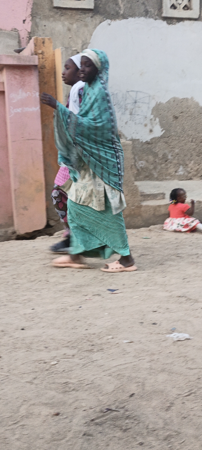 This outdoor image captures a daytime street scene in Rangaza, Nigeria. In the foreground, two young women or girls are walking from left to right on a sandy path. The person in front wears a light teal head covering and a matching long skirt over a lighter top, paired with light peach open-toed sandals. Her dark-skinned face is visible in profile, showing a slight smile. Partially obscured behind her is another dark-skinned individual wearing a white head covering, a light top with a pink pattern, and dark shoes.
On the right side of the mid-ground, a small dark-skinned child sits on the sandy ground or a low step. The child is dressed in a bright red top and a white skirt with a red floral pattern, and has small braids or pigtails. They appear to be looking downwards.
The background consists of weathered walls and structures, suggesting a residential or village setting. On the far left, a light pink column or pillar shows faint, handwritten text, which is mostly illegible but includes fragments resembling "Gulans" and "Bee." The ground is sandy or dirt, with minor debris scattered throughout. The lighting indicates it is daytime, with diffused light and no harsh shadows.