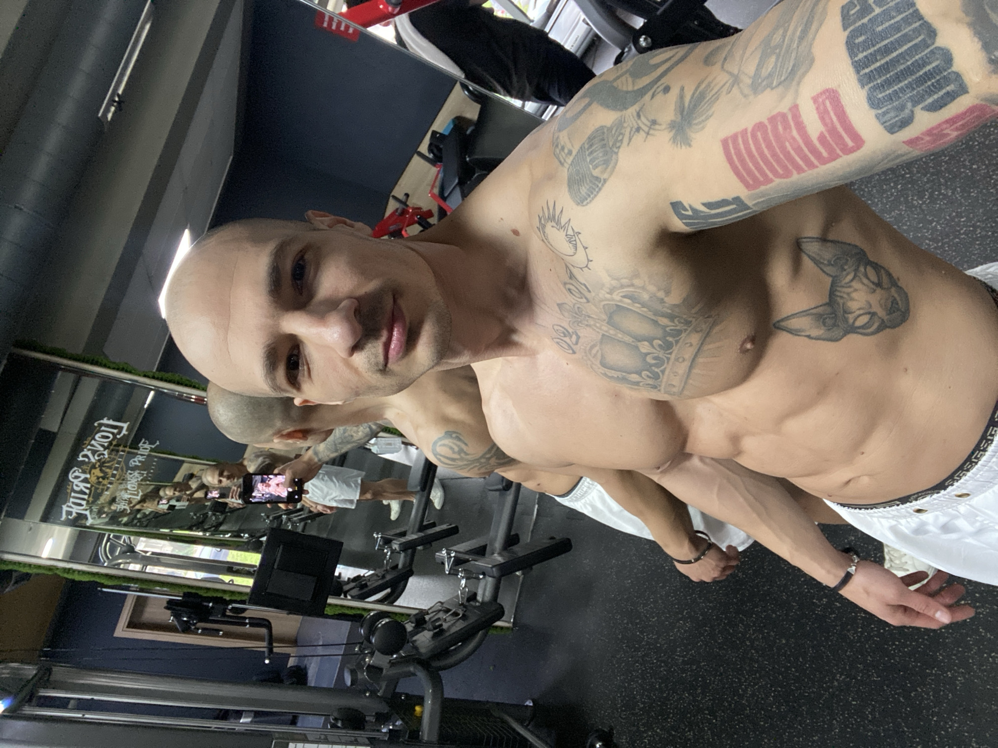 An adult male subject is depicted taking a mirror selfie inside Lion's Pride Gym in Pleven, Bulgaria. The subject is shirtless, bald, and has numerous tattoos covering his chest, abdomen, and arms, including a large crown on his upper chest, a feline head on his lower right ribcage, and text on his left forearm (partially visible "PUMA" in red). He wears white athletic shorts with a dark waistband and a black bracelet on his left wrist. He holds a black smartphone in his right hand, angled towards the mirror to capture his reflection, with the phone screen displaying an image of himself.
The gym environment features dark, speckled rubber flooring, dark blue-grey walls, and sections adorned with vertical artificial green foliage. Various gym equipment, including cable machines with weight stacks and benches, are visible in the background and reflections. A large mirror covers a significant portion of the wall, reflecting the subject, gym equipment, and a light brown wooden door. The gym's logo, "LION'S PRIDE GYM" with a lion's head graphic, is visible as a reflection on the mirror. Overhead lighting fixtures are also reflected.