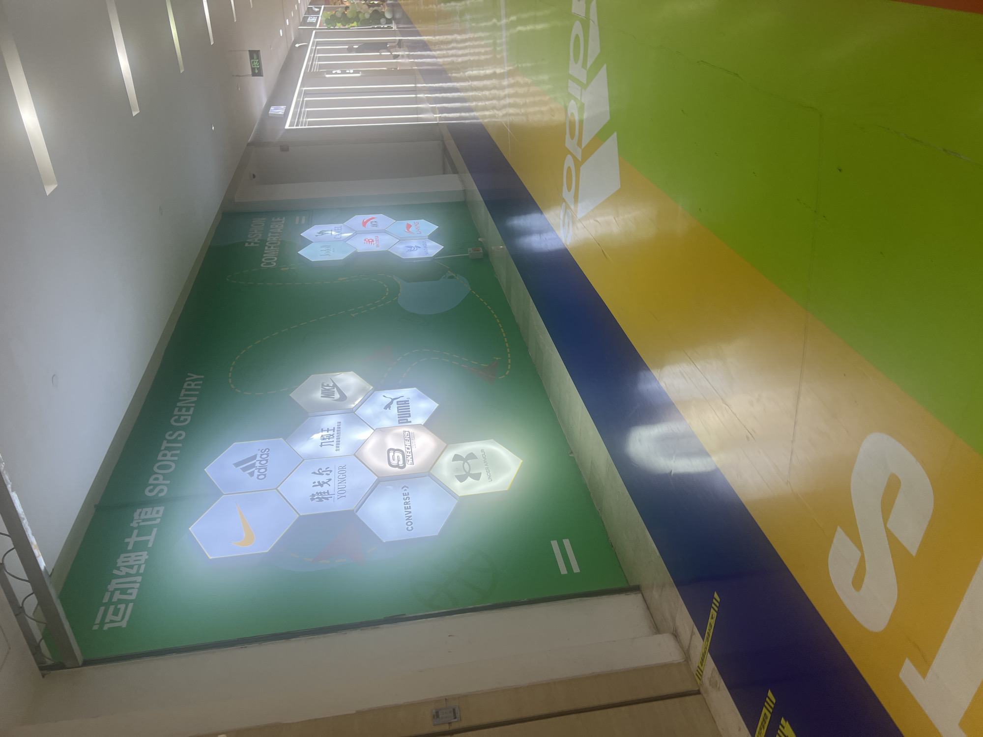 The image shows an interior view of a shopping mall or commercial space, likely within Liandu District, China, featuring a large green wall mural with hexagonal displays of various sportswear brands. The brands visible include Nike, Adidas, Youngor, Converse, Skechers, Puma, and Under Armour. Text in Chinese and English reading "运动细士馆 SPORTS GENTRY" is present on the wall. Below the mural, the floor is decorated with bold stripes of blue, yellow, and green, with large white "adidas" lettering and the Adidas logo visible on the yellow section. The overall environment appears clean and well-lit, suggesting a commercial area designed for sports apparel retail. There are no people clearly visible in the immediate foreground or engaging in any specific actions. In the background, a few figures can be seen walking in the distance, and store entrances with illuminated vertical lines are present.
